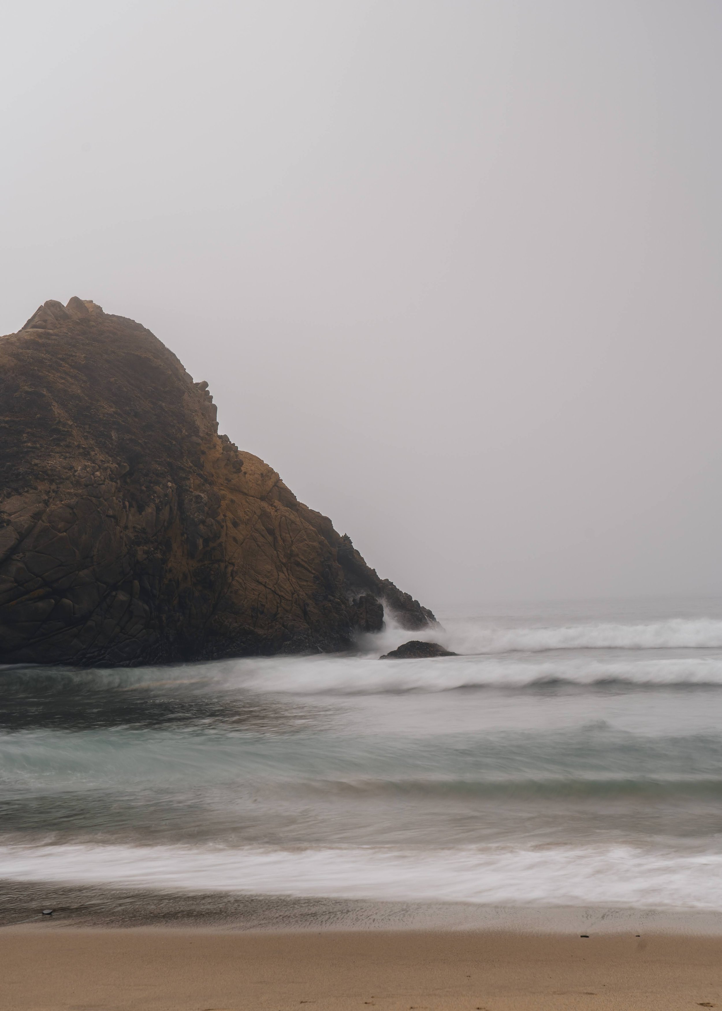 A rocky coastline with waves crashing against the shore and a foggy sky.