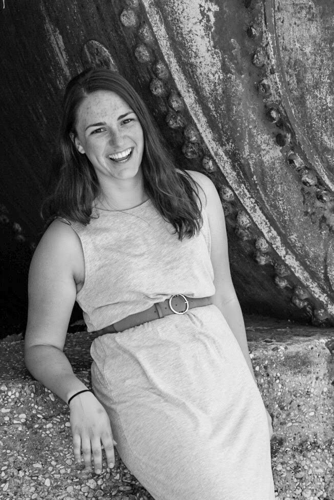 A woman with shoulder-length hair smiling and wearing a sleeveless dress with a belt, sitting against a rocky background.