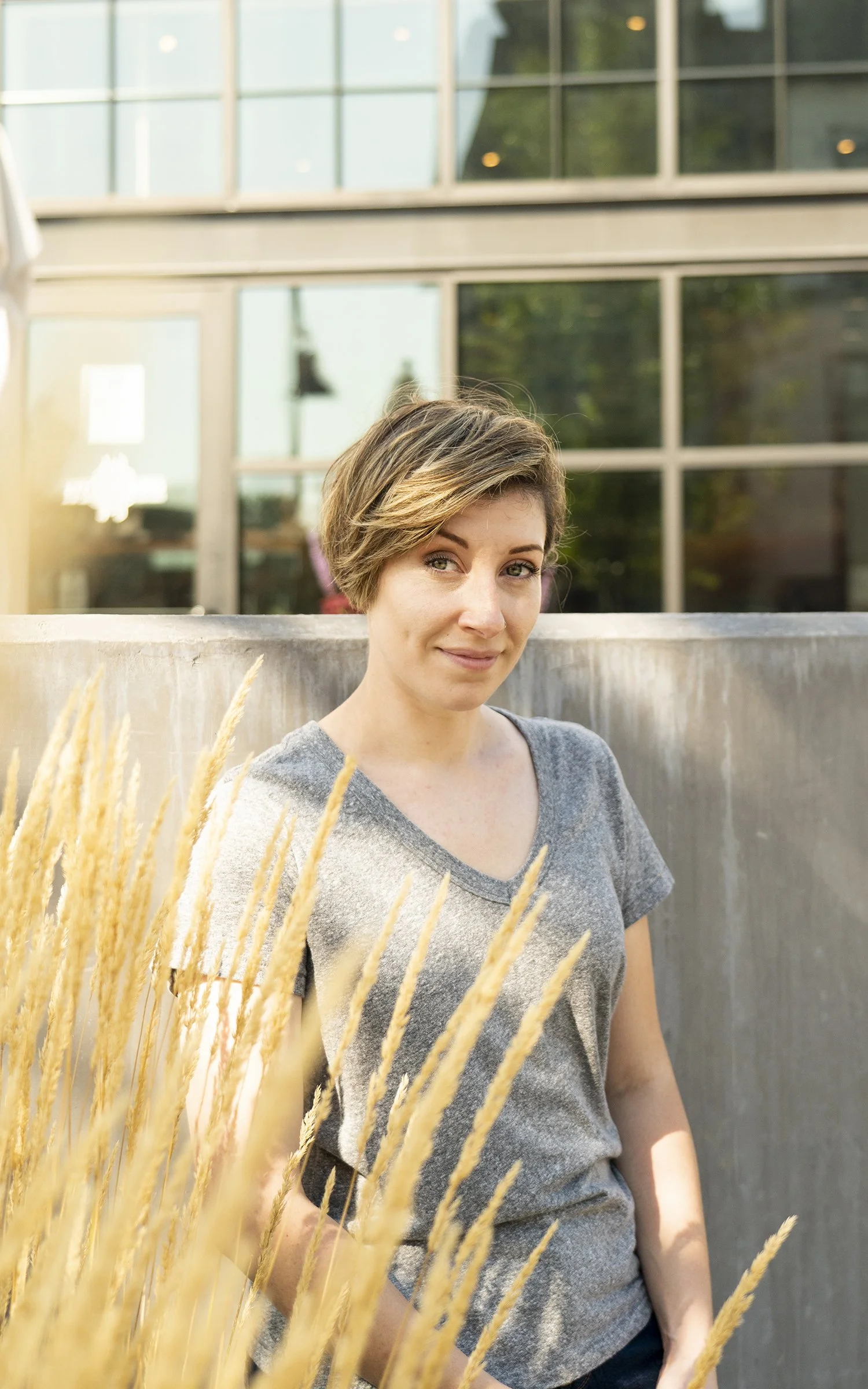 A woman with short, brown hair and a gray T-shirt standing outdoors in front of a modern glass building, partially obscured by tall, golden grass in the foreground.