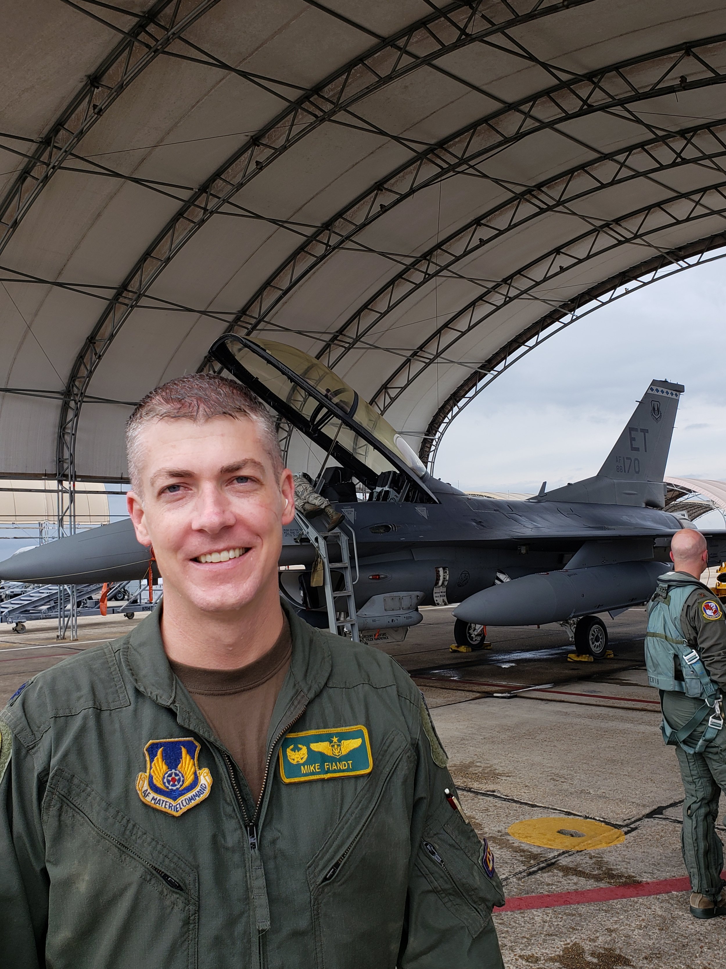 Mike “Hondo” Fiandt, owner of Hondo’s Wood Co, in a military flight suit standing in front of an F-16 fighter jet.