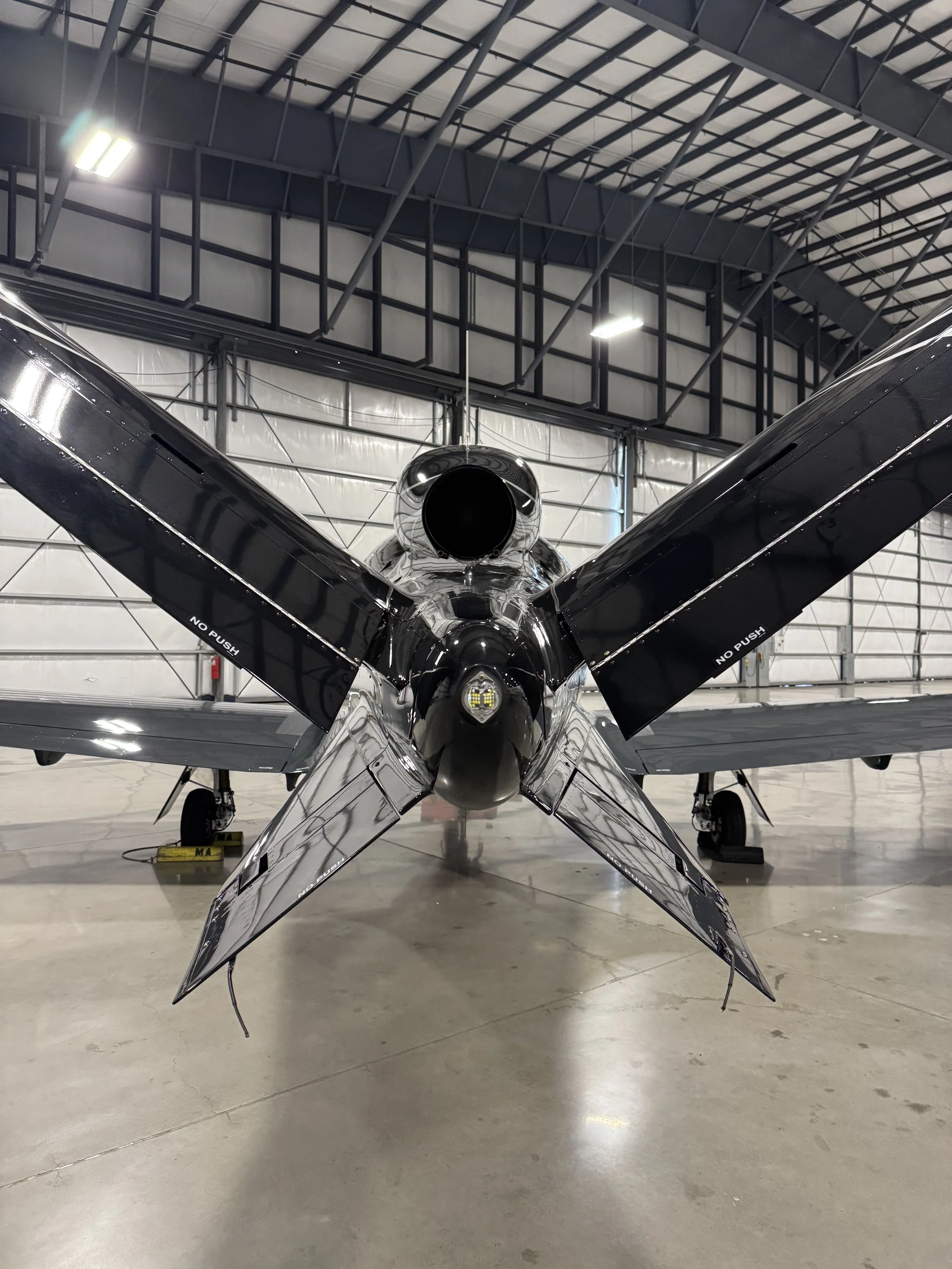 A small, shiny, black airplane inside an aircraft hangar with a high ceiling, metal beams, and bright overhead lighting.