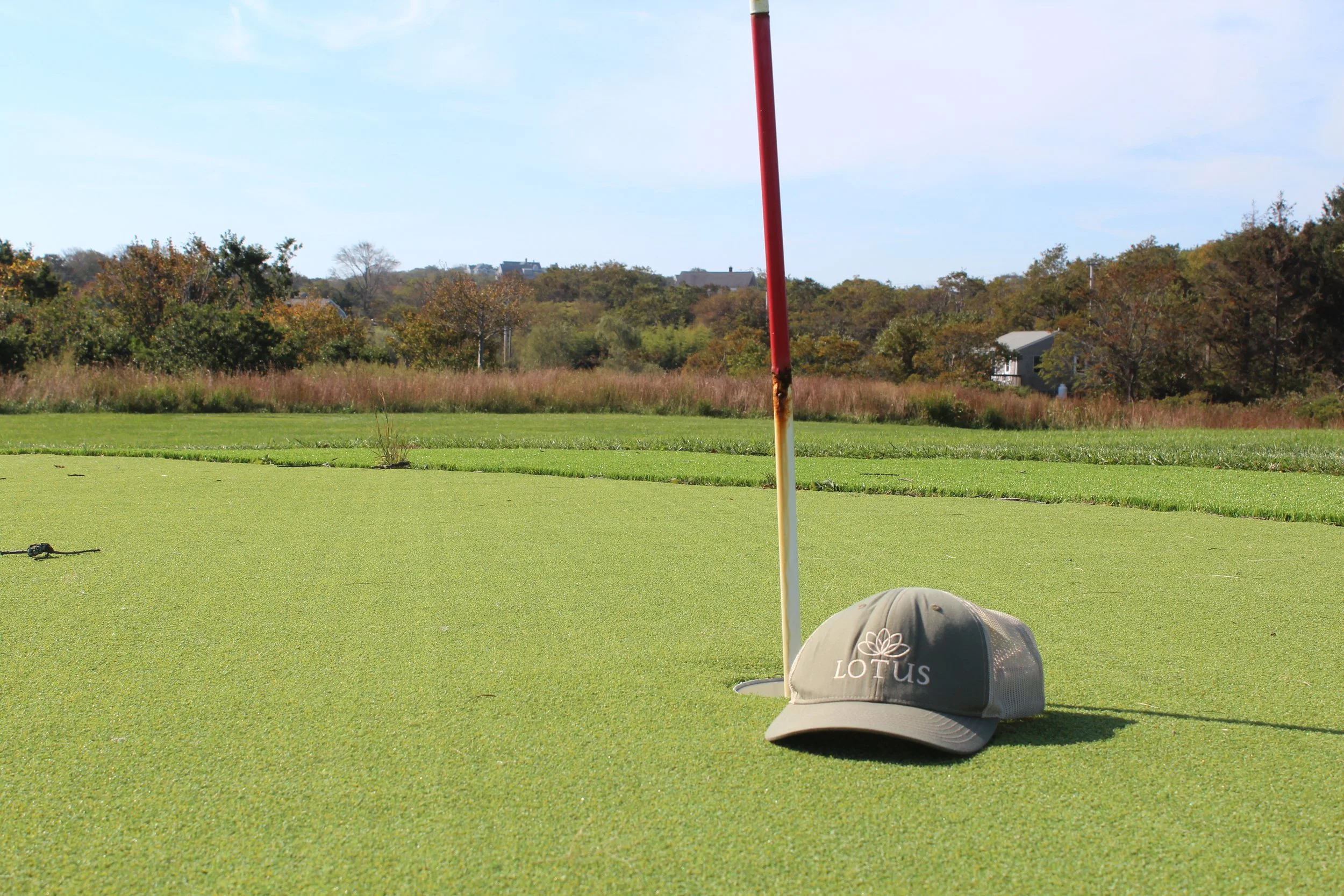 Golf course with a putting green, a red flagstick, and a fitted cap with lotus logo on the ground.