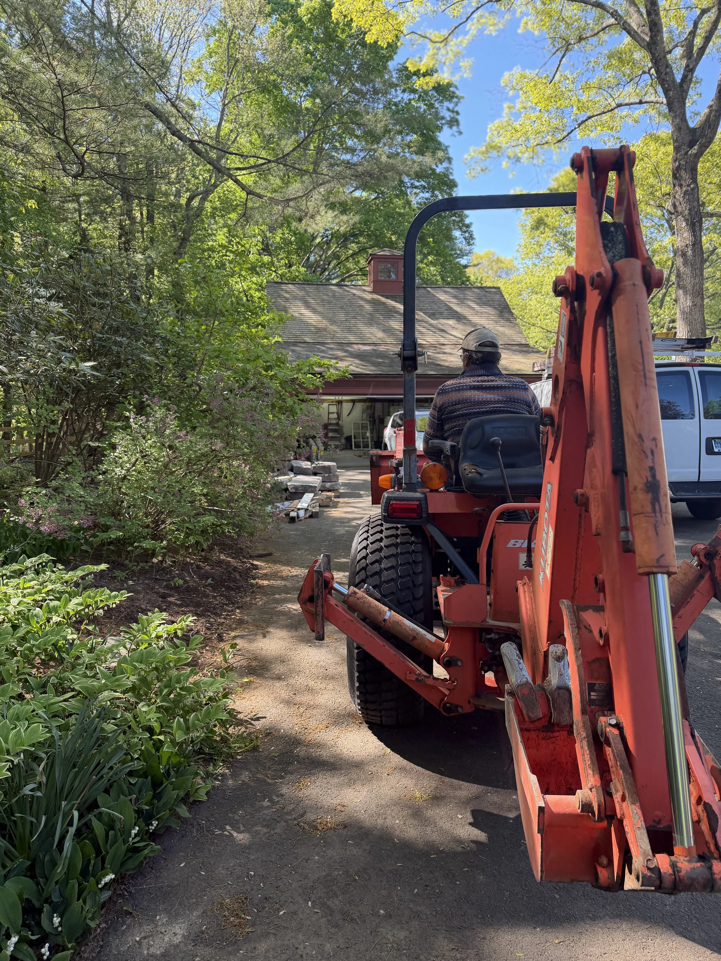 Person operating a small orange excavator on a dirt driveway in front of a house with trees and greenery on a sunny day.