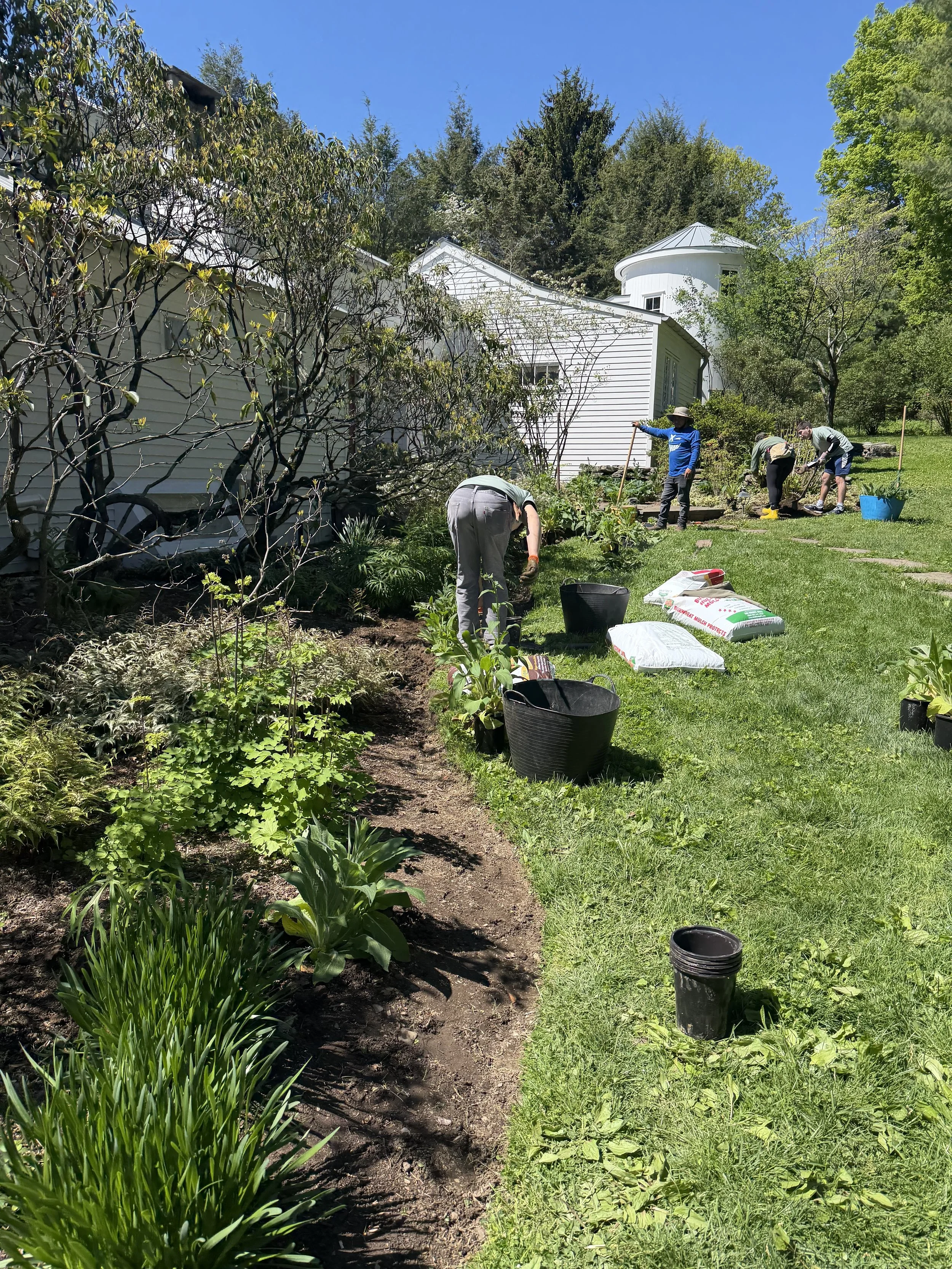 People planting in a garden near a white house with a turret, surrounded by trees under a clear blue sky.