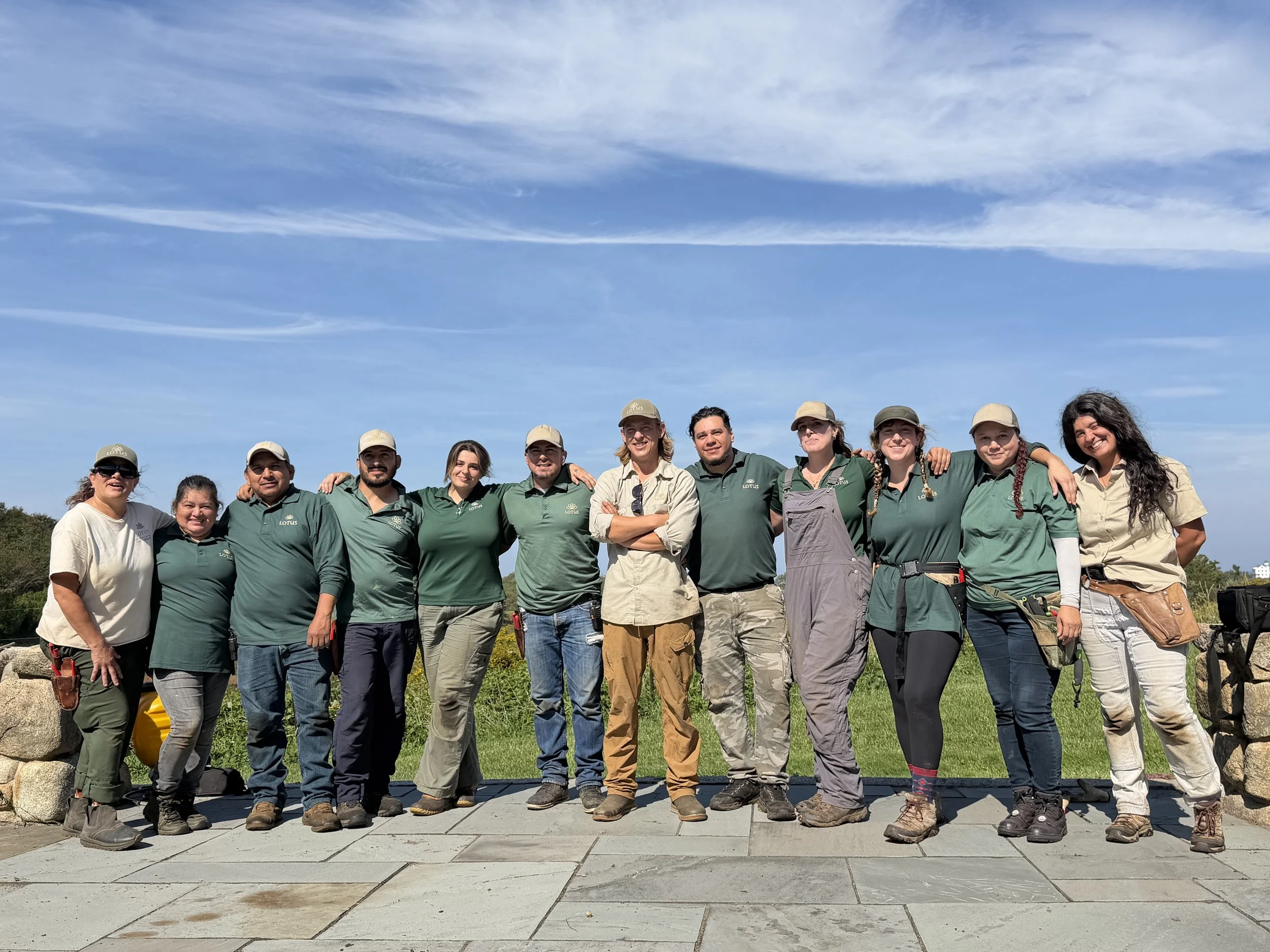 Group of people standing together outdoors under a partly cloudy sky, some wearing green shirts with a logo, smiling for the photo.