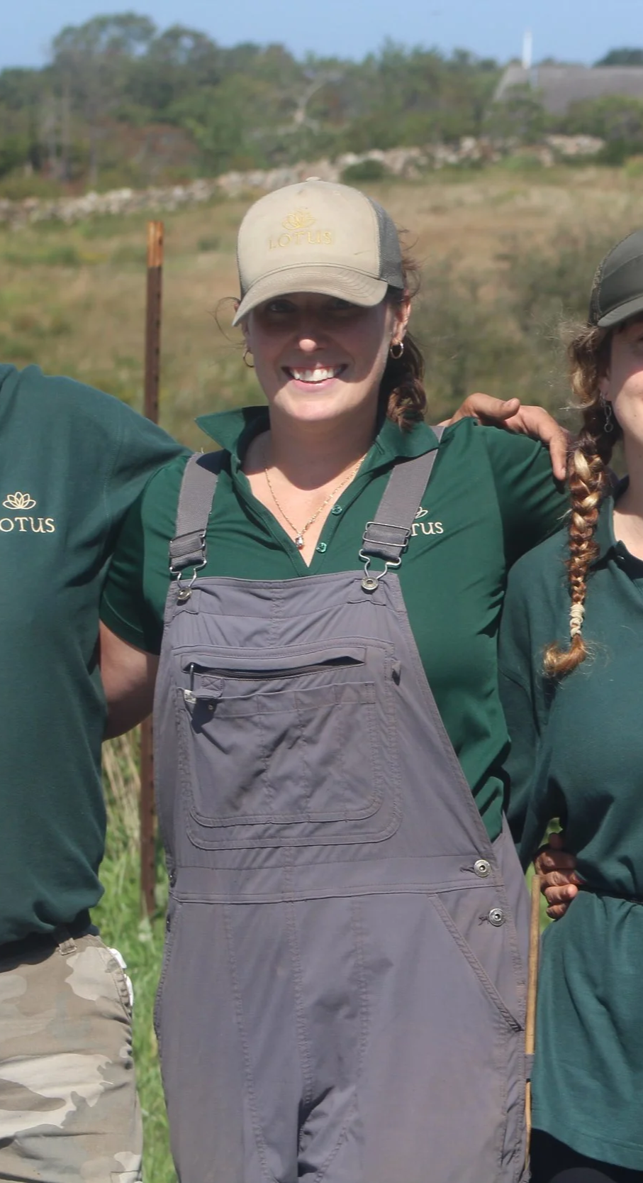 A woman wearing a beige Lotus cap, green shirt with Lotus logo, and gray overalls stands outdoors, smiling with her arm around someone next to her.
