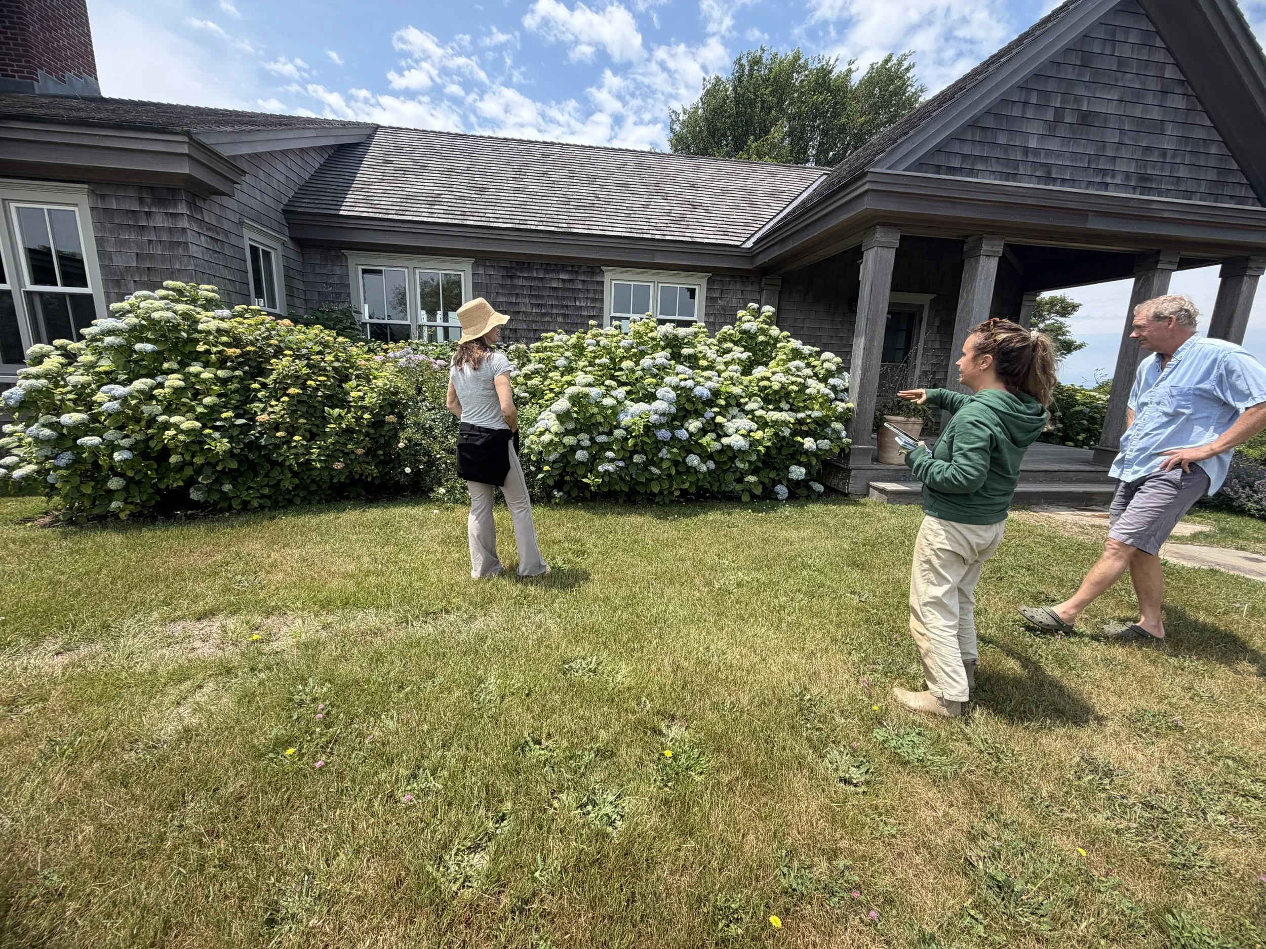 Four people standing outside a house with a porch, talking and looking at flowers, under a partly cloudy sky.