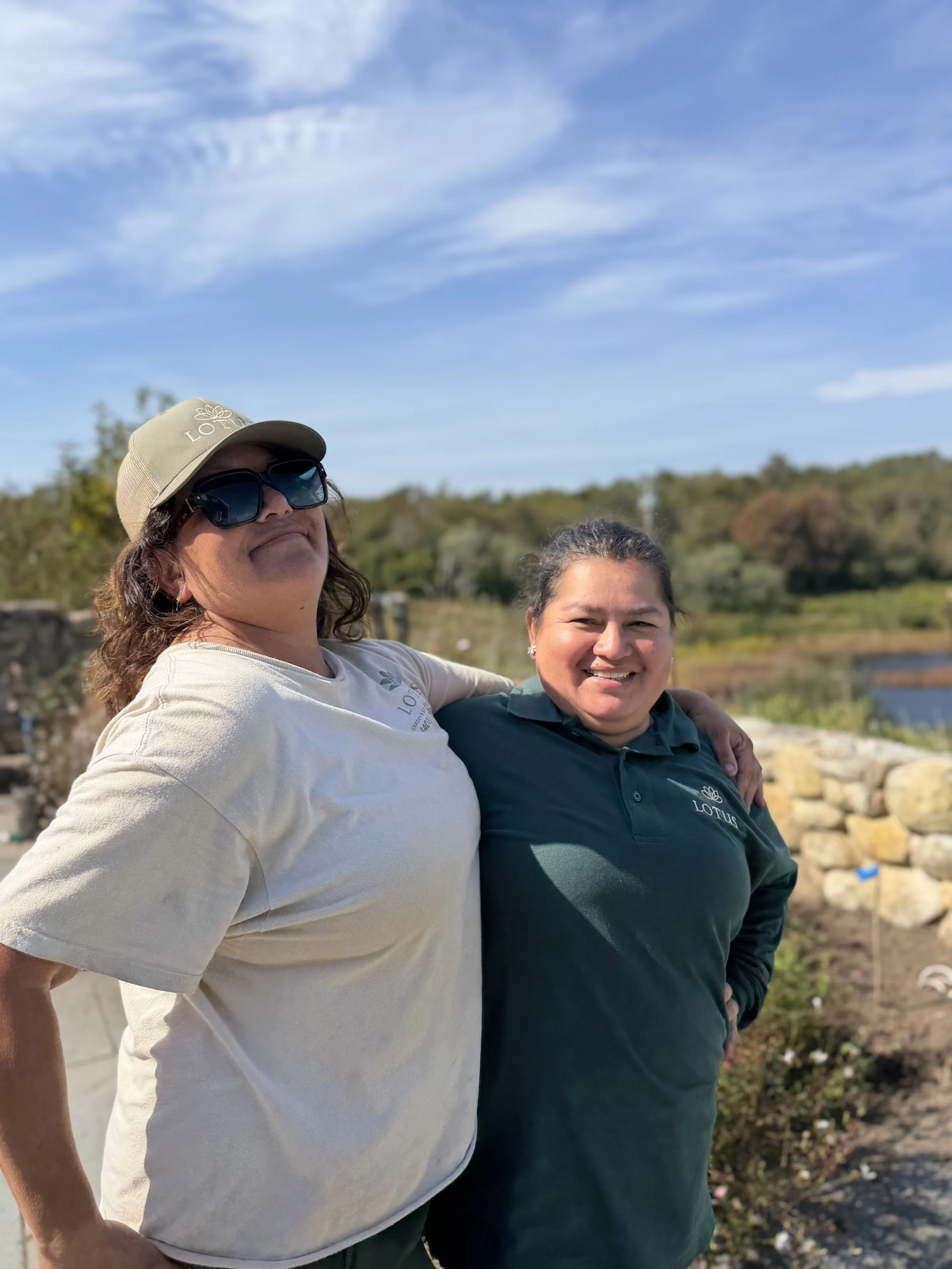 Two women standing outdoors in front of a pond and greenery, smiling, with one woman in a beige cap and sunglasses, the other in a dark green shirt with a Lotus logo.