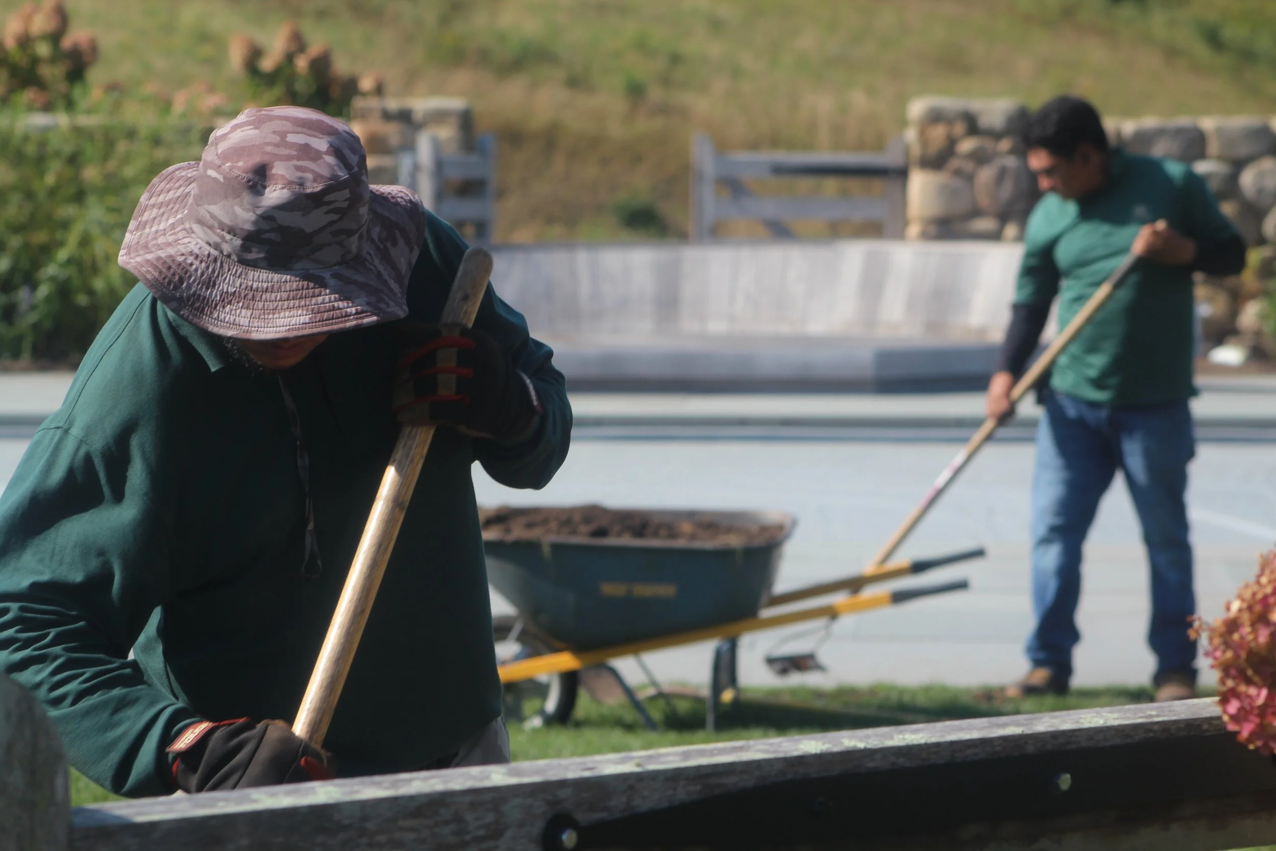 Two people raking leaves in an outdoor area during daytime. One person is wearing a camouflage sun hat and a green jacket, while the other is dressed in a green shirt. There are wheelbarrows and a backdrop of a hillside and stone wall.