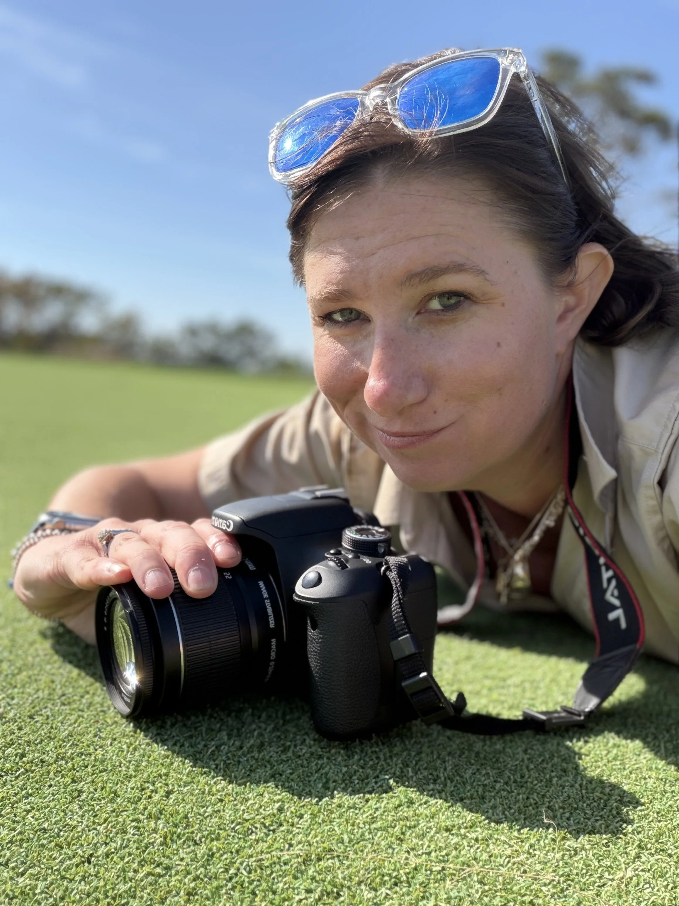 Woman lying on green grass with a camera, wearing sunglasses on her head, on a sunny day with blue sky and some trees in the background.