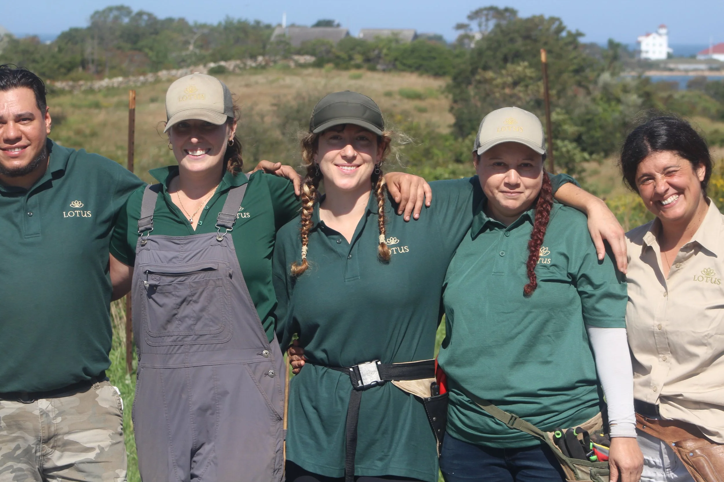 Group of five people standing outdoors, smiling, with arms around each other, wearing green shirts and hats with a Lotus logo, in a natural setting with grassy hills and trees in the background.