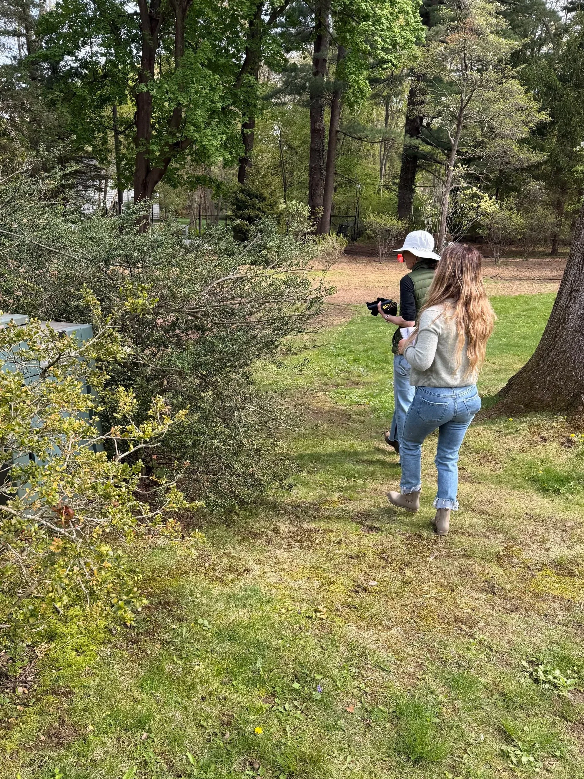 Two women walking outdoors on a grassy area near trees and bushes. One woman is holding a clipboard, wearing a hat, and the other has long wavy hair and is wearing a gray sweater and boots.