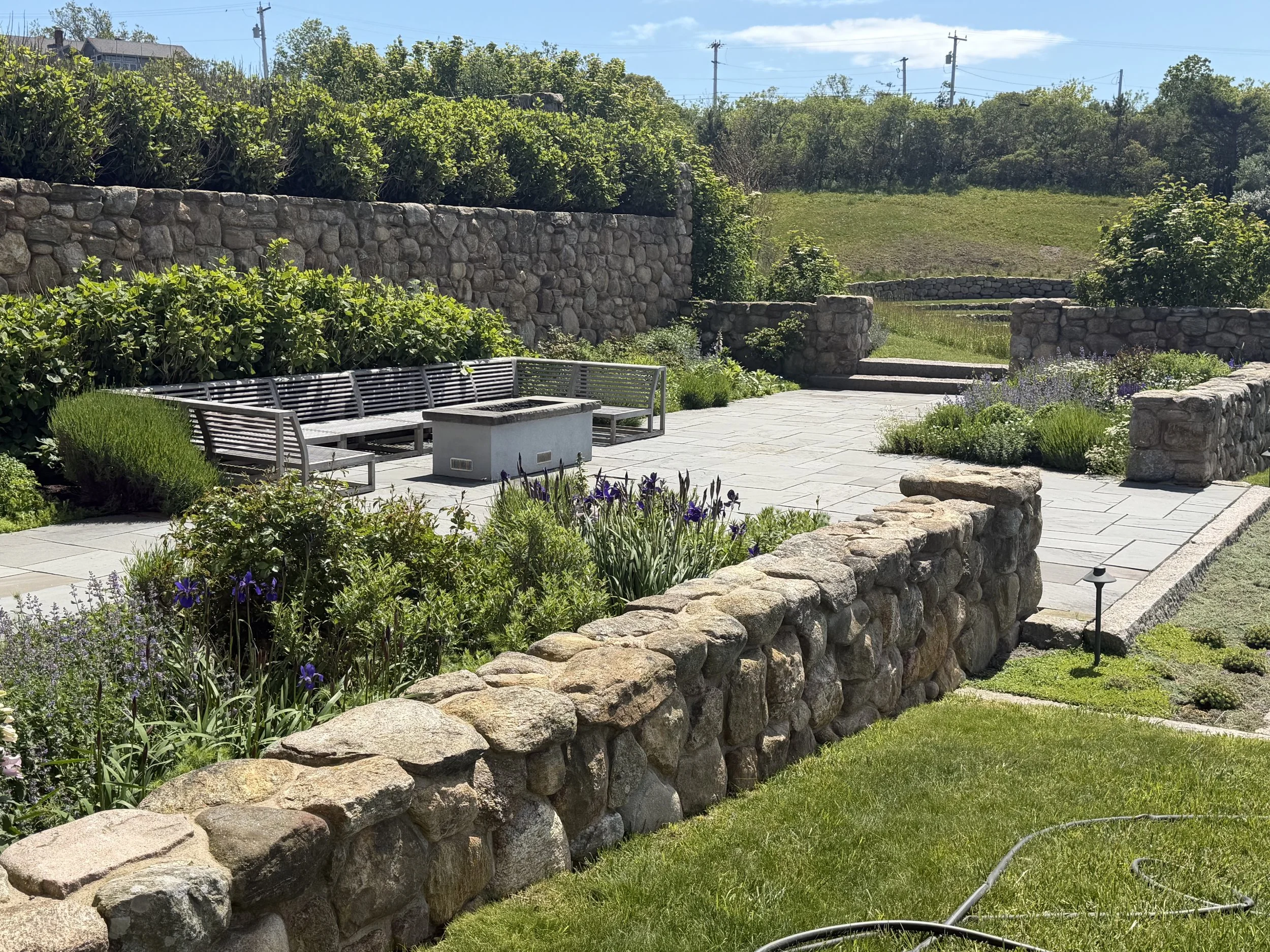 A landscaped backyard with stone retaining walls, lush green plants, purple flowers, and a patio with outdoor seating. In the background, there are electrical poles, trees, and a grassy hillside under a cloudy sky.