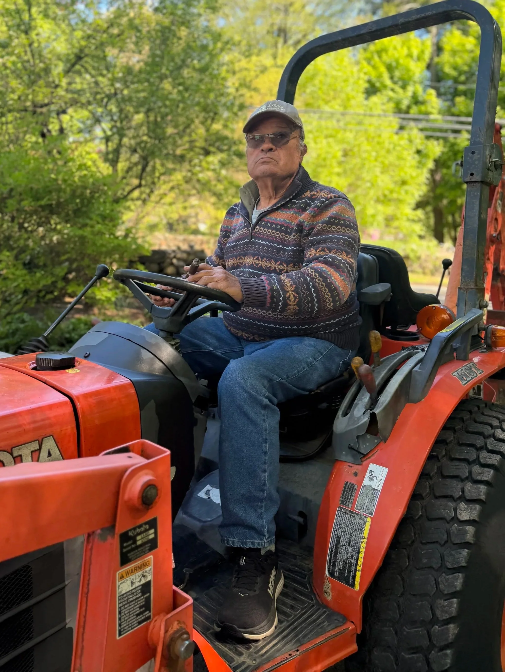 Older man with sunglasses and a cap riding a Kubota orange tractor through a lush green outdoor area with trees in the background.