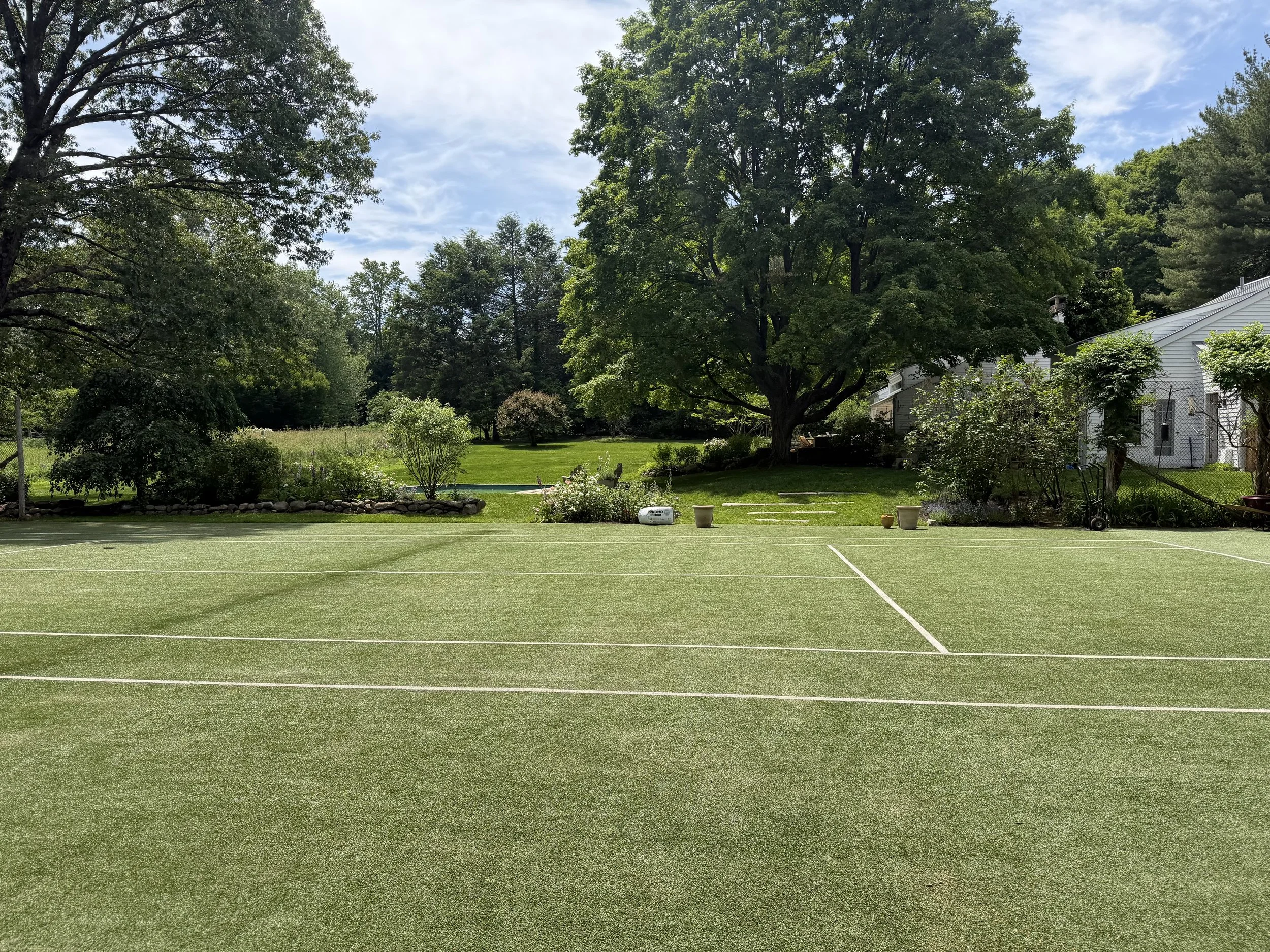 An outdoor tennis court surrounded by lush green trees and a house with a white exterior.
