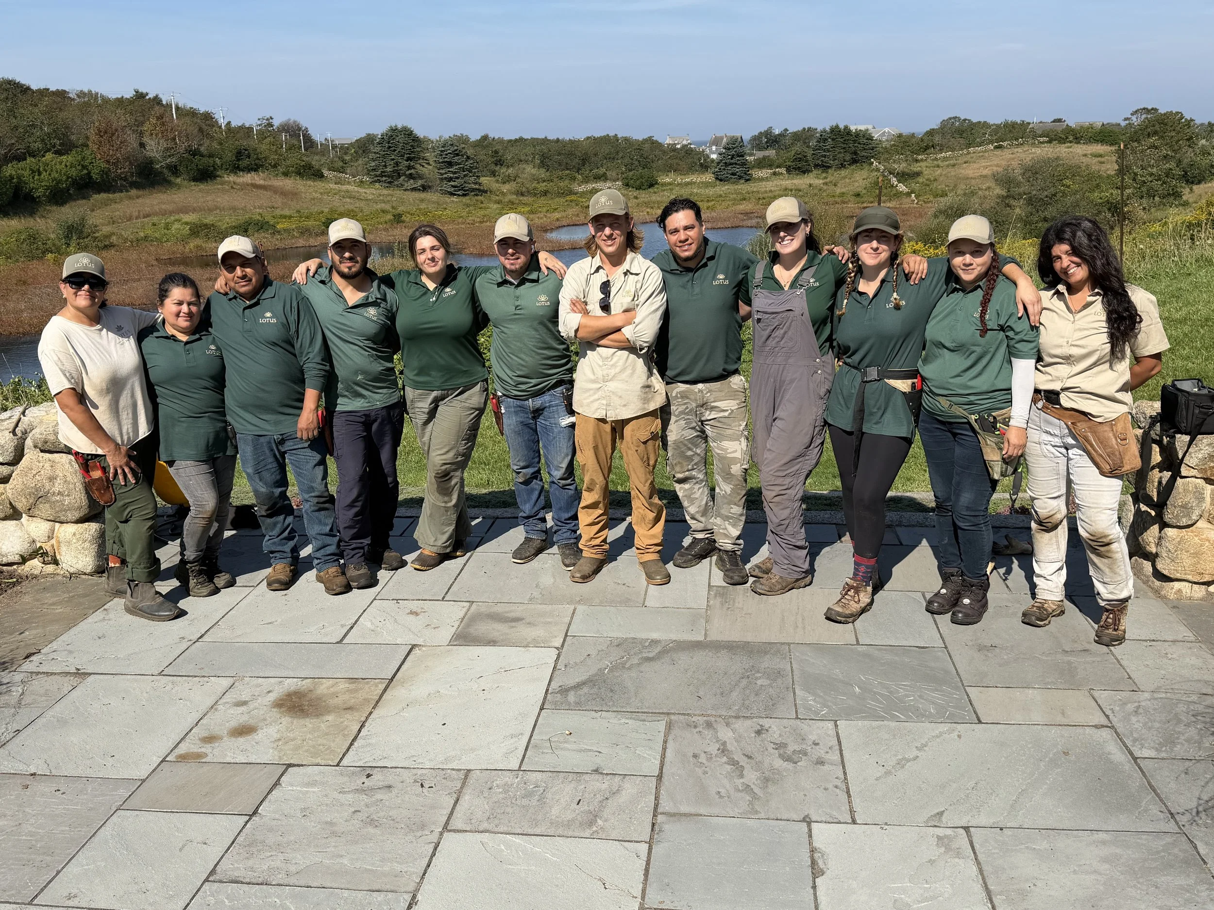 A group of eleven people standing outdoors on a stone pathway with a scenic landscape of trees, grass, and water behind them. They are smiling and have their arms around each other, dressed casually with some wearing hats and uniforms.