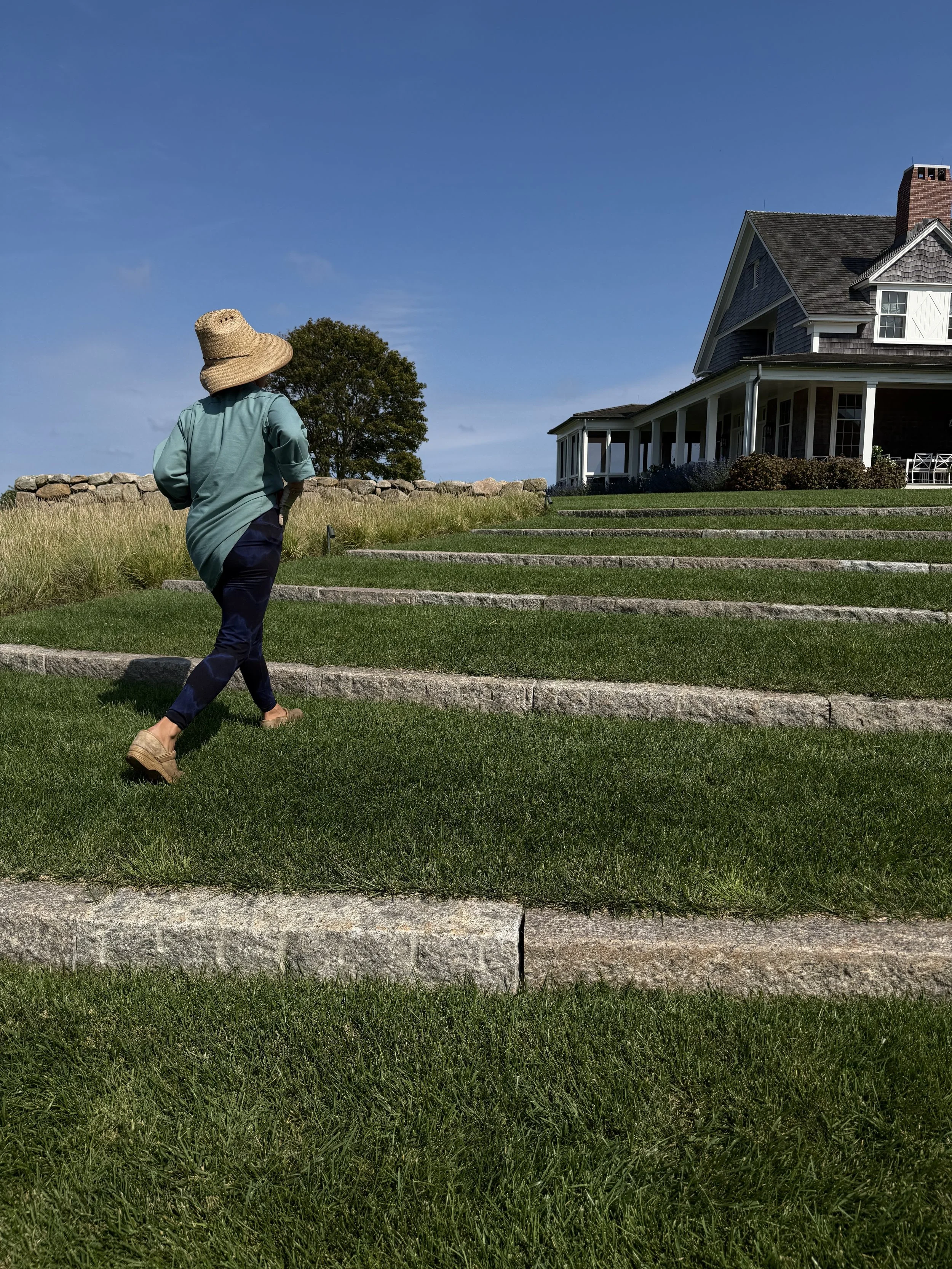 A woman with a large sun hat and teal shirt walking on stone steps in front of a large house with a porch and green lawn.