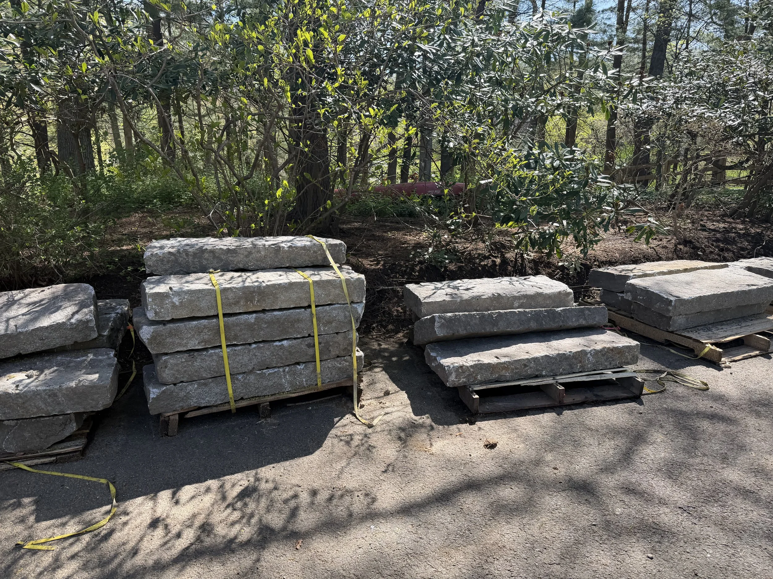 Stacks of concrete pavers with yellow straps in front of a wooded area with trees and shrubs.