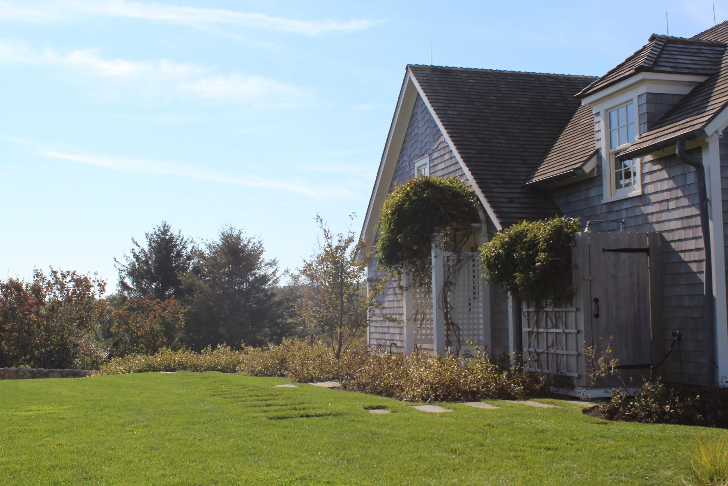 The image shows a house with a wooden shingle exterior and dormer windows, surrounded by a lush green lawn and trees, under a clear blue sky with some clouds.