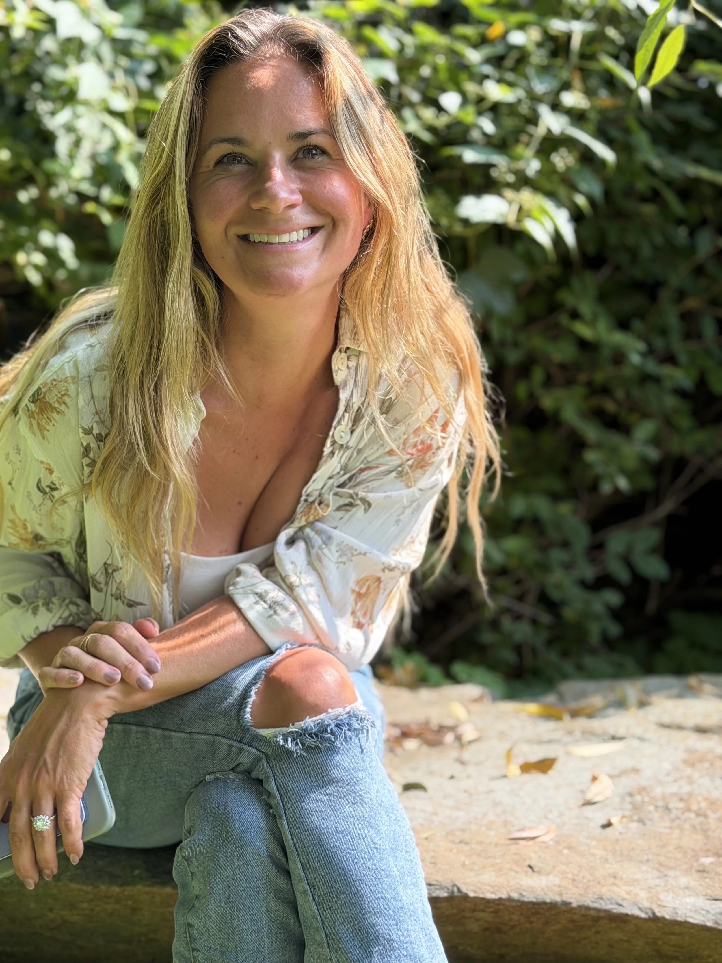 A woman smiling outdoors with her arm around another person, with greenery in the background and sunlight highlighting her hair.