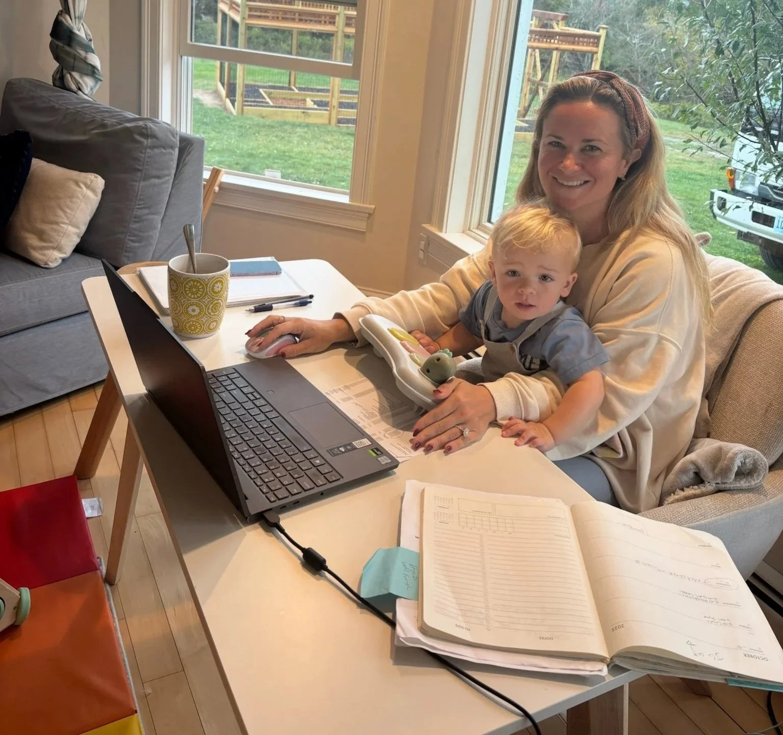 A woman and a young boy sitting at a white table in a bright room with large windows, working on a laptop and paperwork, with a backyard visible outside.