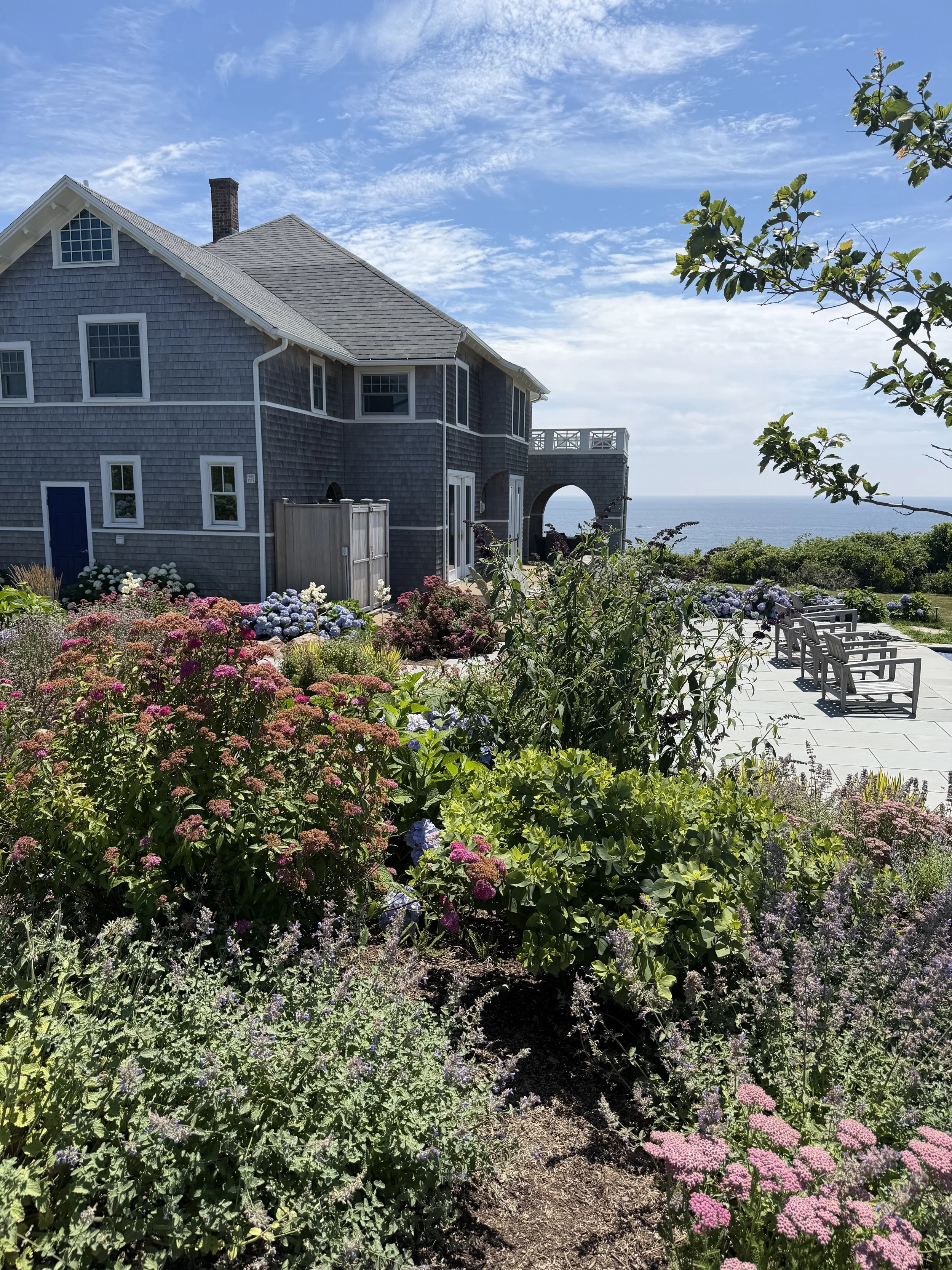 A house by the ocean with a garden filled with colorful flowers, a patio with chairs, and a scenic view of the sea and blue sky.