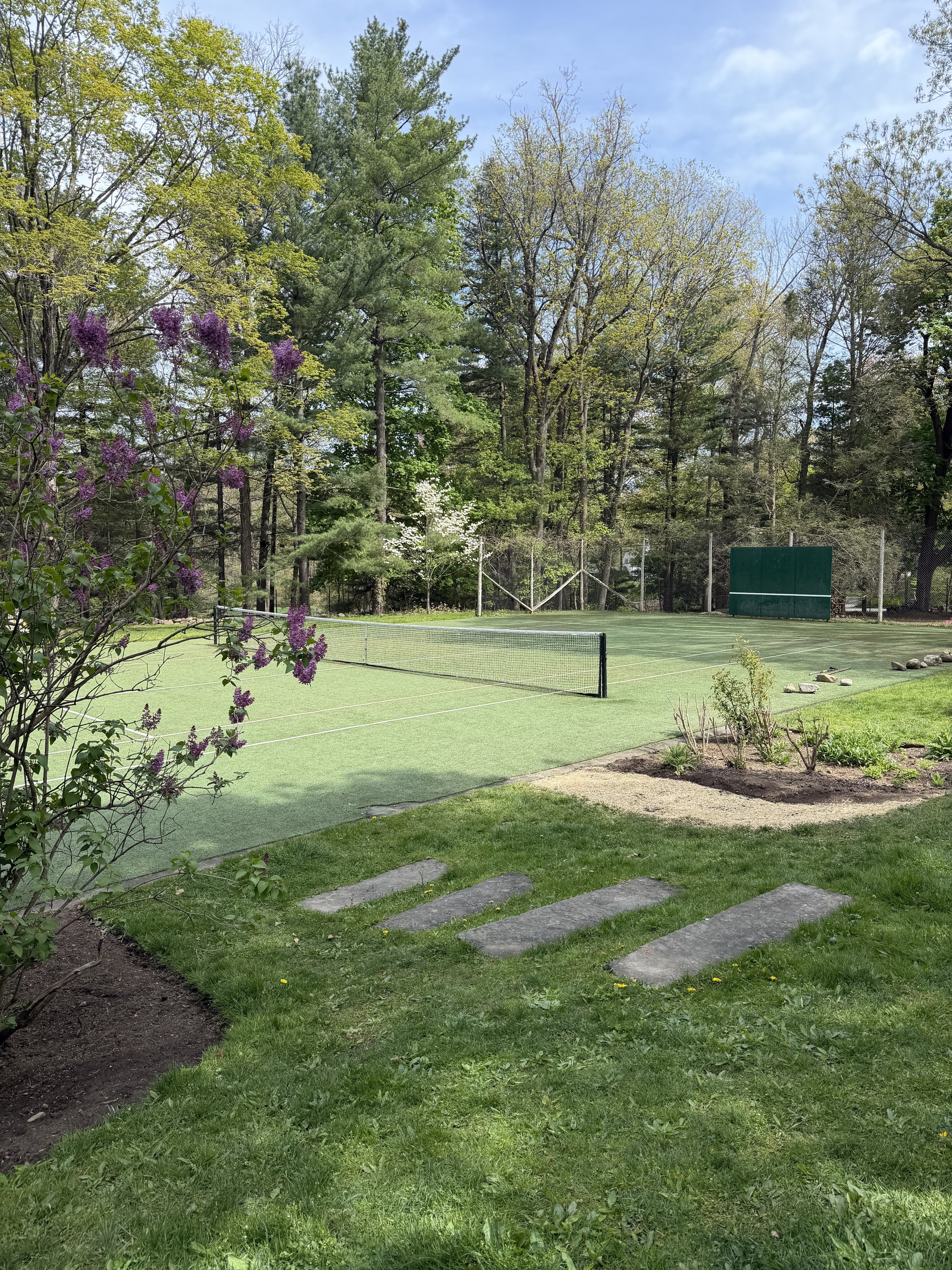 A green tennis court surrounded by trees and flowering shrubs, with a fence and a backstop at one end, and steps leading up from a grassy area in the foreground.
