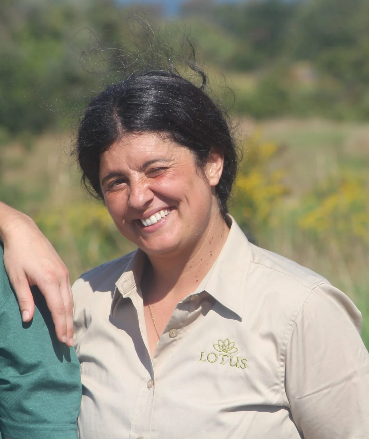 Close-up of a woman with dark hair smiling and winking, wearing a beige Lotus shirt, outdoors with blurred green background.