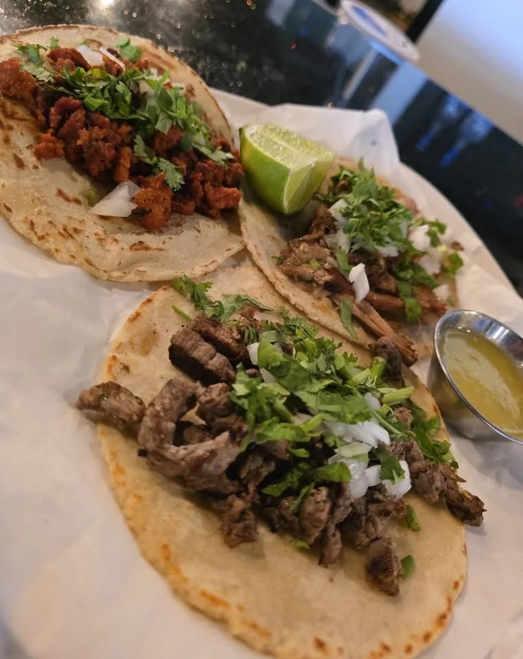 Three tacos with different fillings, lime wedge, and a small container of yellow sauce on a white paper-lined tray.