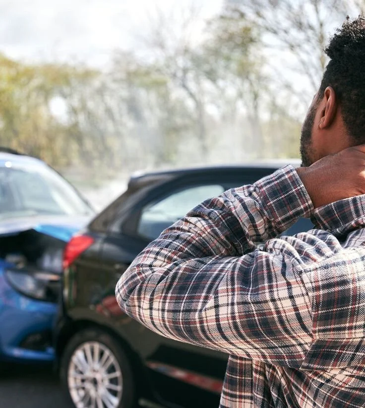 Man in plaid shirt holding his neck in front of two parked cars after a car accident.