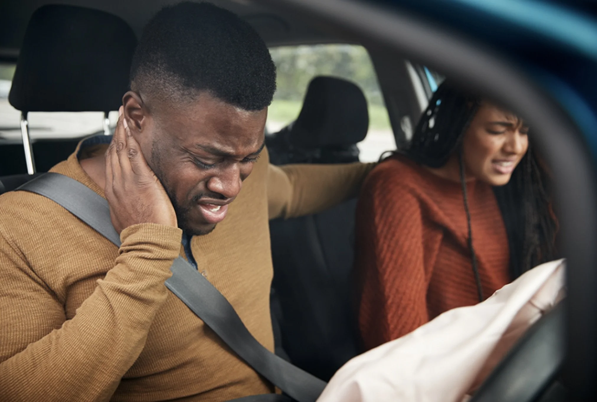 A man and a woman in the back seat of a car, both appear to be in pain or distress, with the man holding his neck and the woman grimacing.