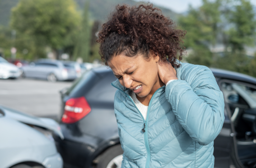 A woman with curly hair wearing a blue jacket standing outdoors near parked cars, holding her neck and looking down with a pained expression.