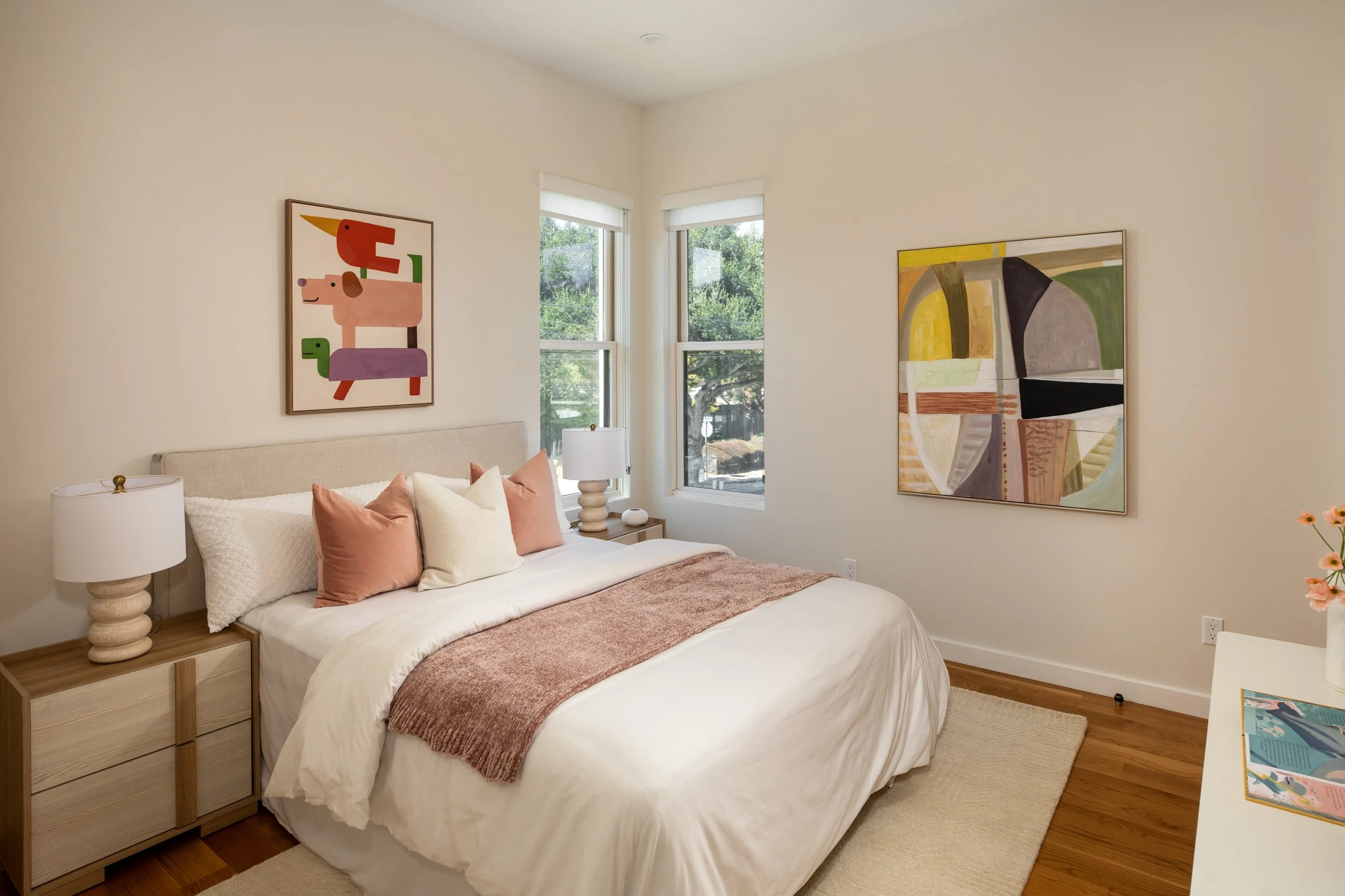 A bedroom with a bed covered in white linens and pink pillows, two bedside lamps, two large windows with views of trees, colorful abstract artwork on the walls, and a small table with books.