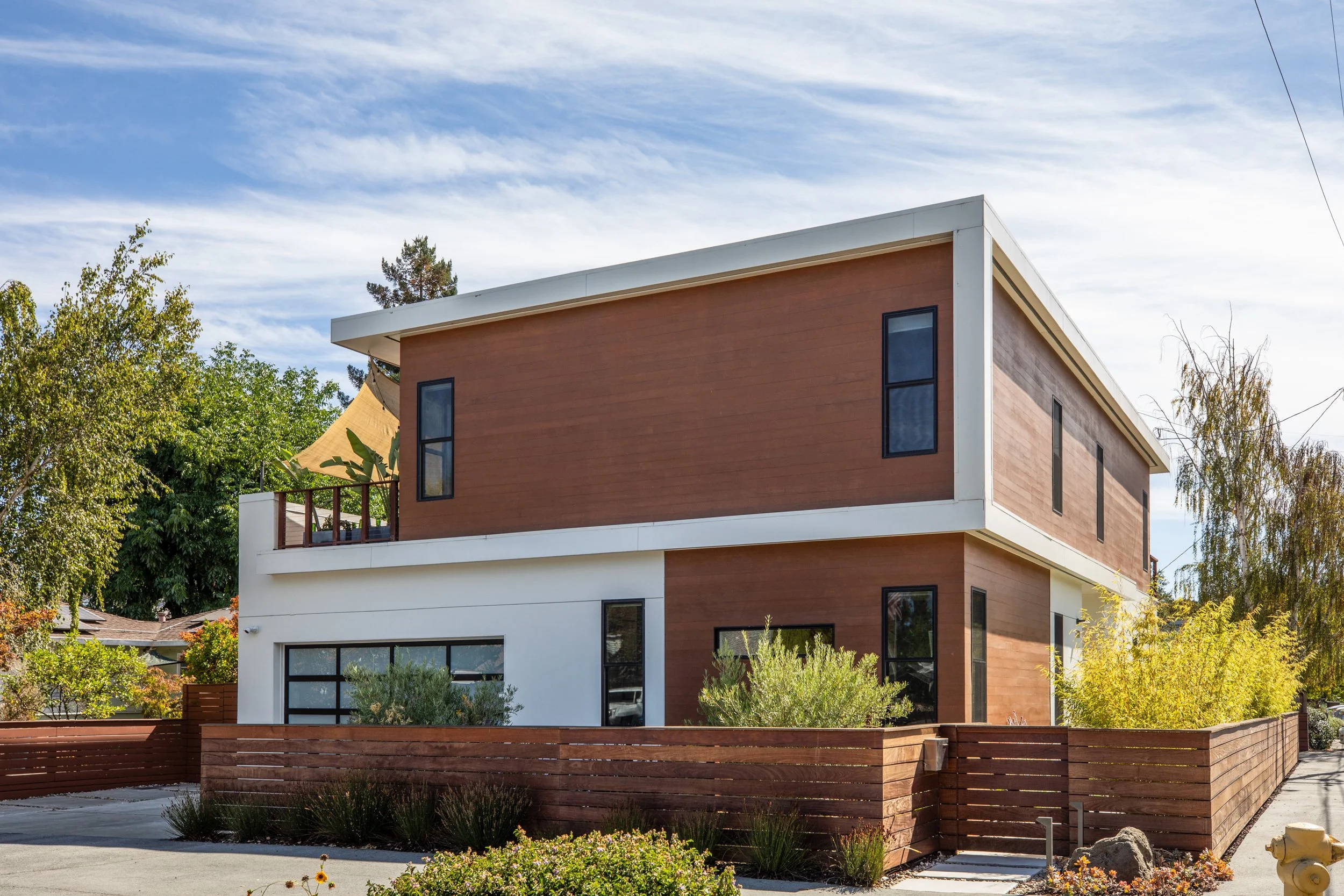 Modern multi-story house with a combination of white and brown exterior walls, large windows, and a wooden fence. The house is set against a backdrop of trees and a partly cloudy sky.