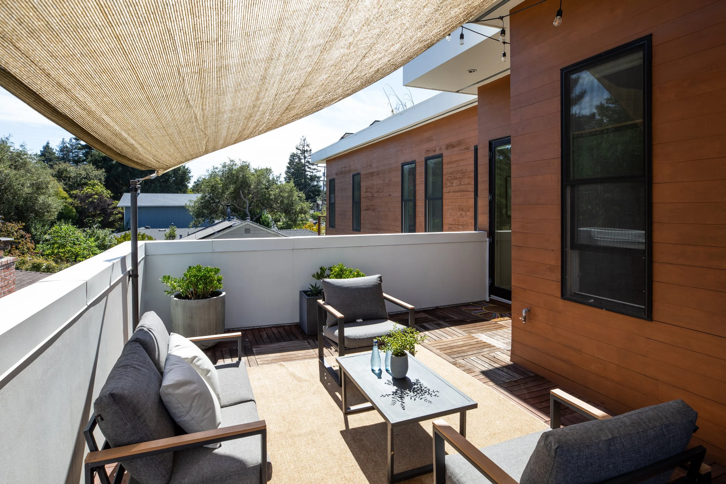 An outdoor balcony with cushioned chairs, small tables, potted plants, and a shade sail covering part of the space, with a view of trees and neighboring houses.