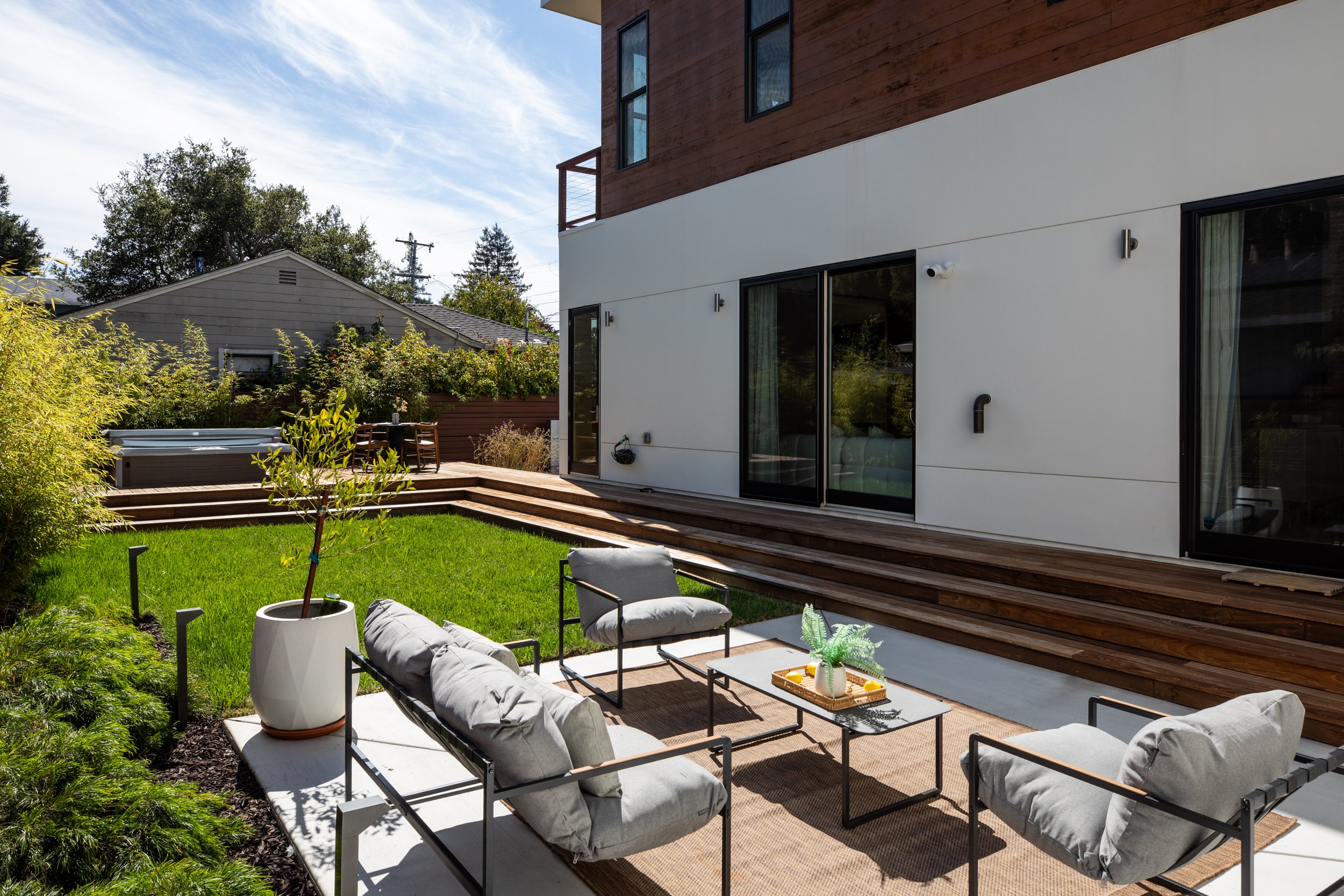 A backyard patio with gray cushioned chairs and a small rectangular black table, a potted plant on the table, a tan outdoor rug, surrounded by green bushes and plants, with a modern house featuring large glass sliding doors and wooden accents in the 