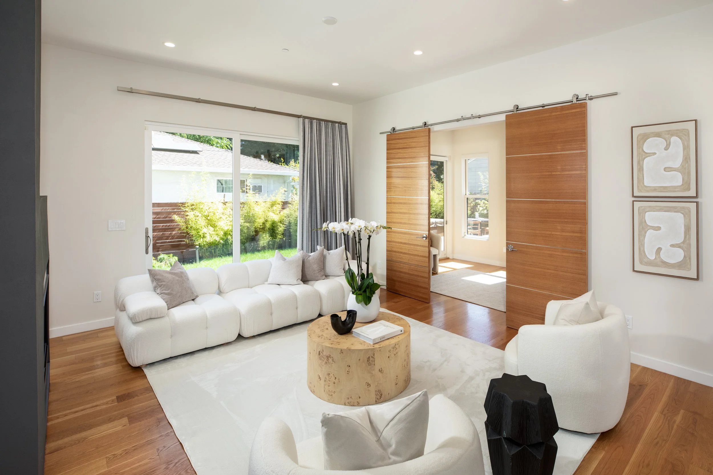 Living room with white sofa, armchair, round coffee table, large windows with curtains, artwork on wall, and sliding wooden doors.
