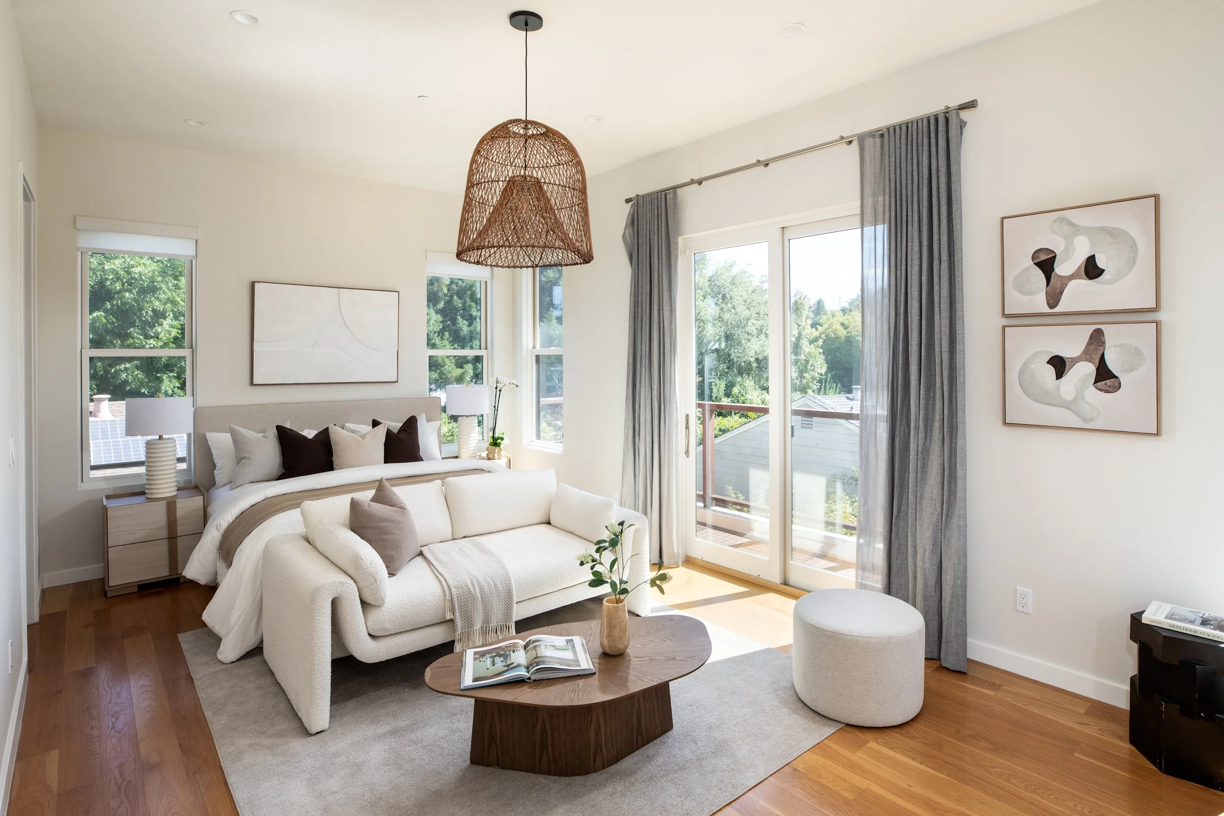 Bright modern living room with large windows, white sofa, wooden coffee table, abstract art on the wall, and a wicker pendant light.