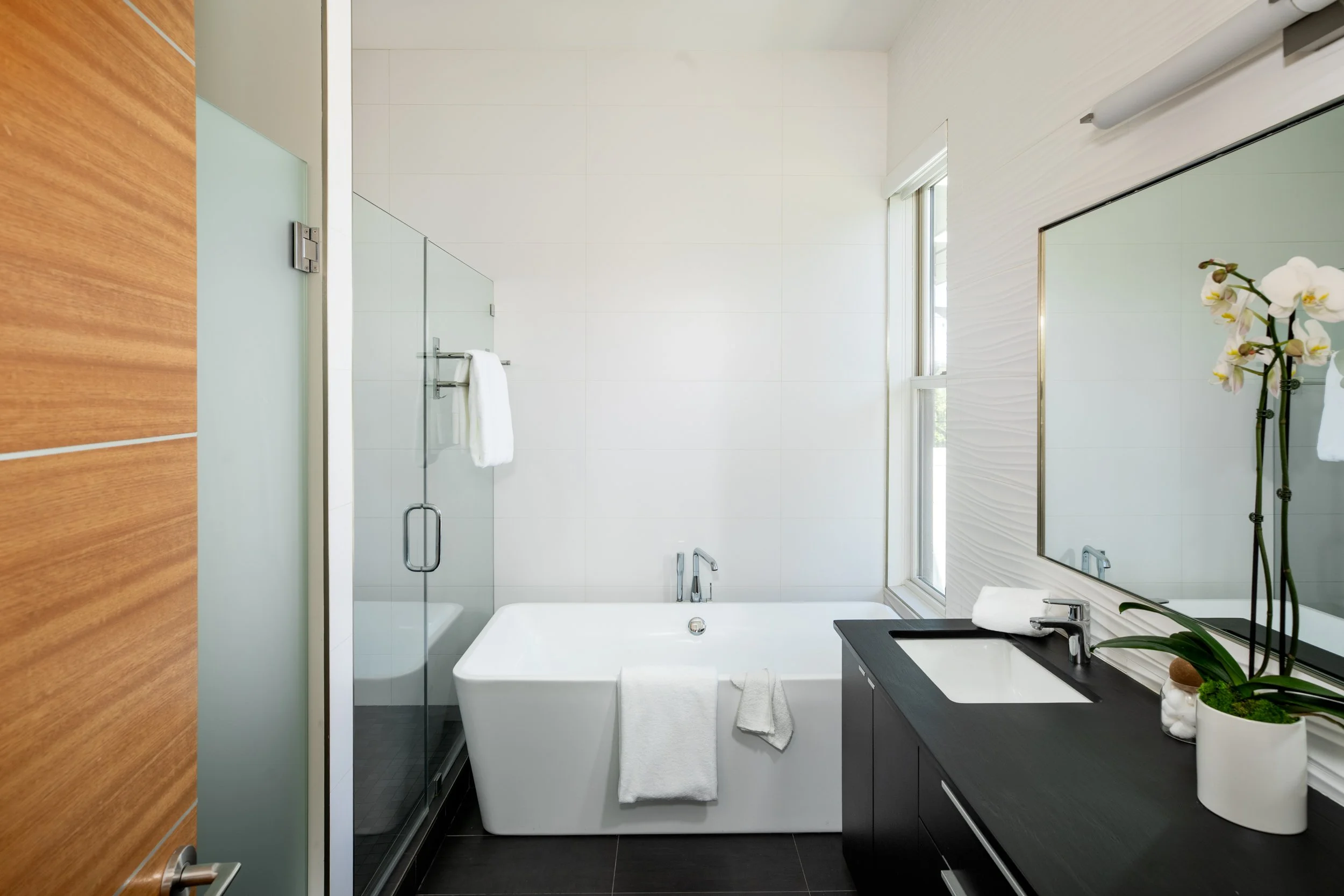 Modern bathroom with a white bathtub, a black vanity with a sink, a large mirror, and a window with natural light. Potted orchid on the vanity.