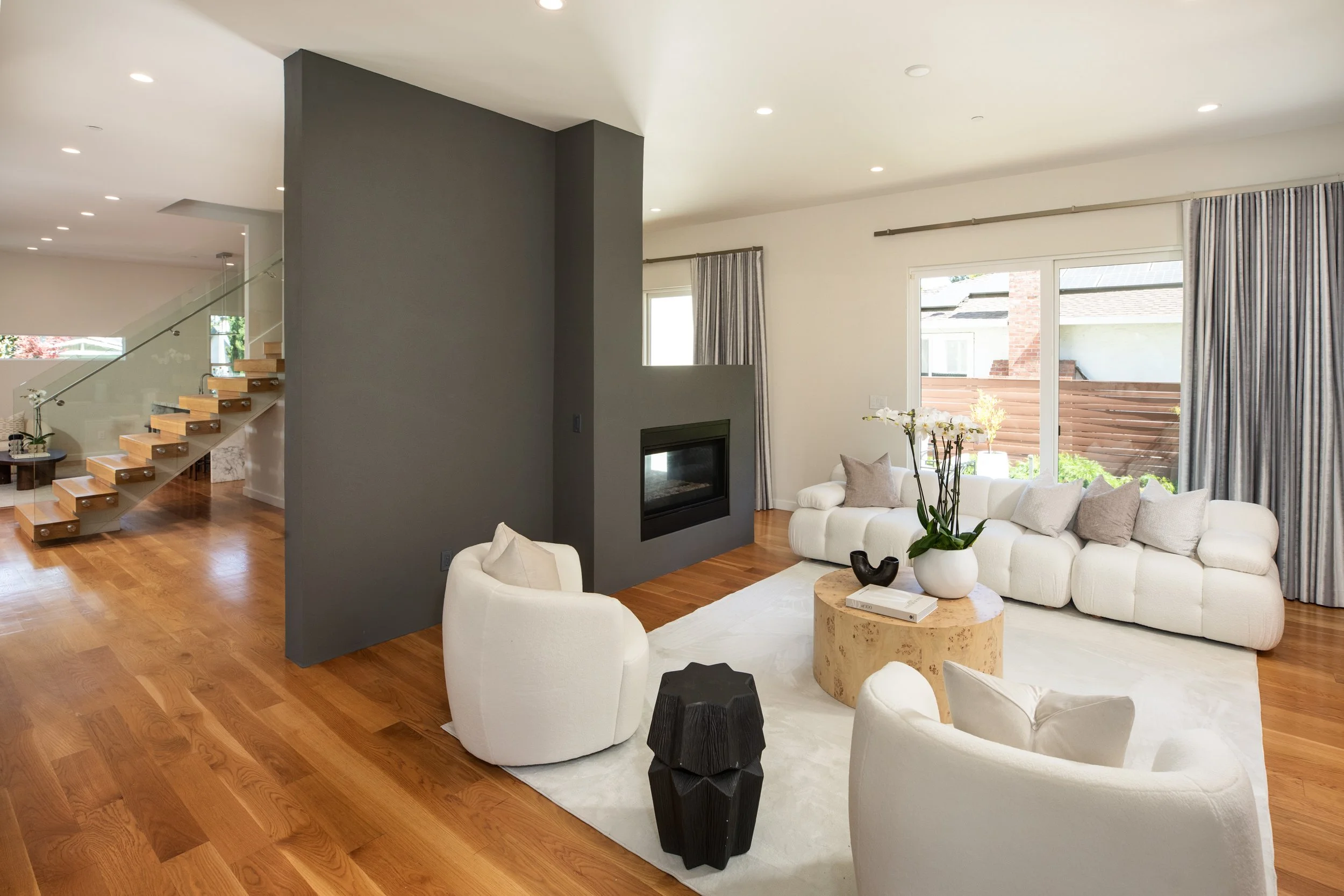 Living room with white sofa and chairs, wooden coffee table with plant, gray accent wall with fireplace, hardwood floors, large windows with curtains, staircase in background.
