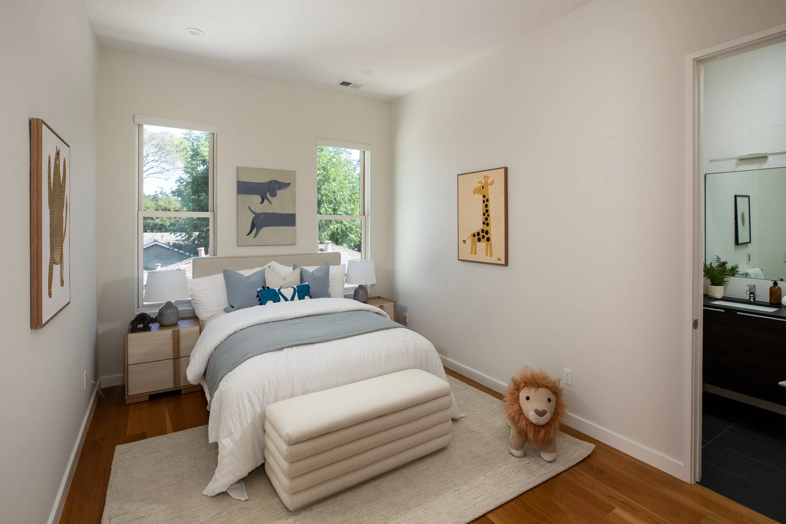 A bedroom with white walls, a bed with white bedding and blue pillows, artwork of animals on the walls, a lion plush toy on a cream rug, and a window showing trees outside.