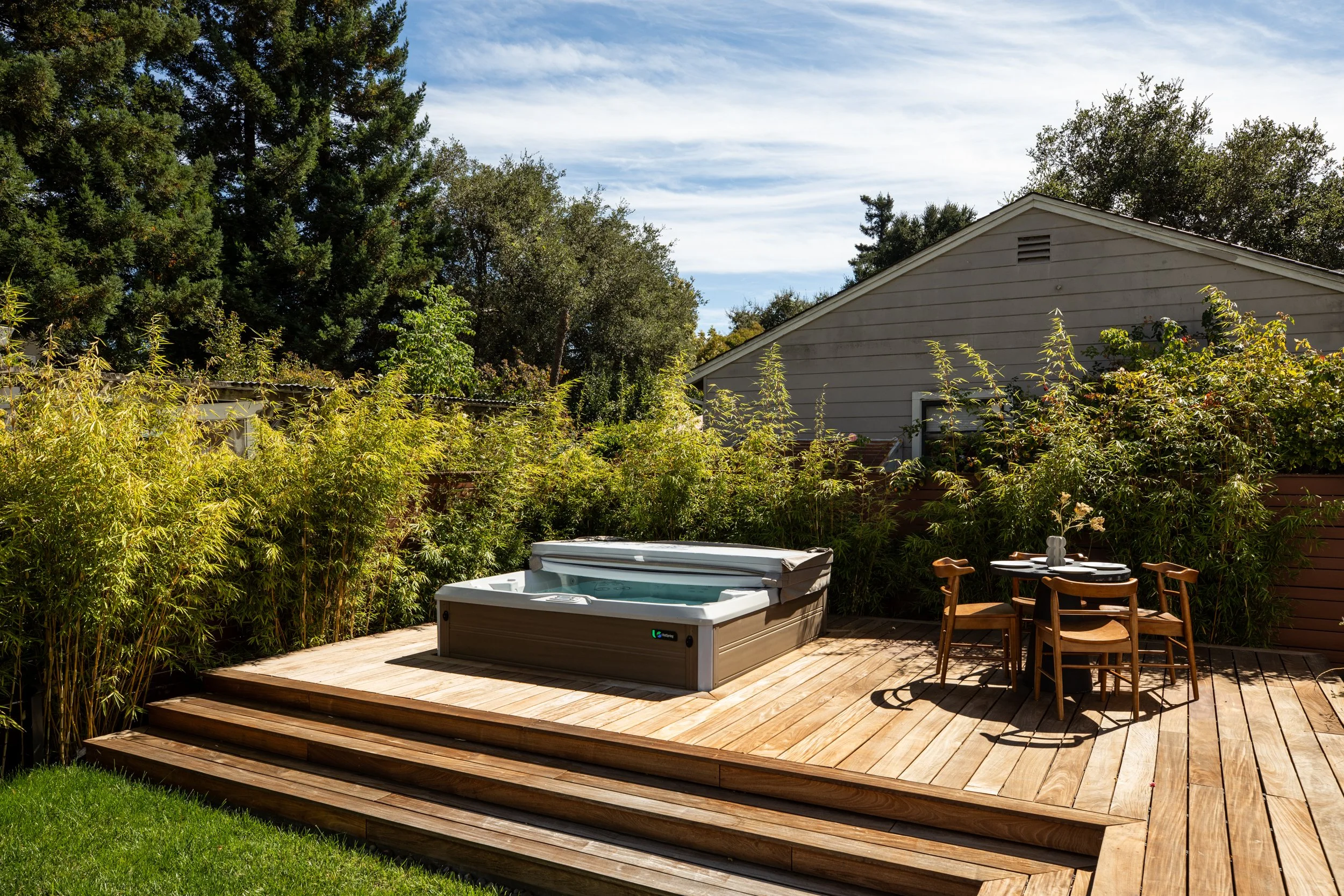 Backyard patio with hot tub on wooden deck, surrounded by greenery, with outdoor chairs and a house in the background.