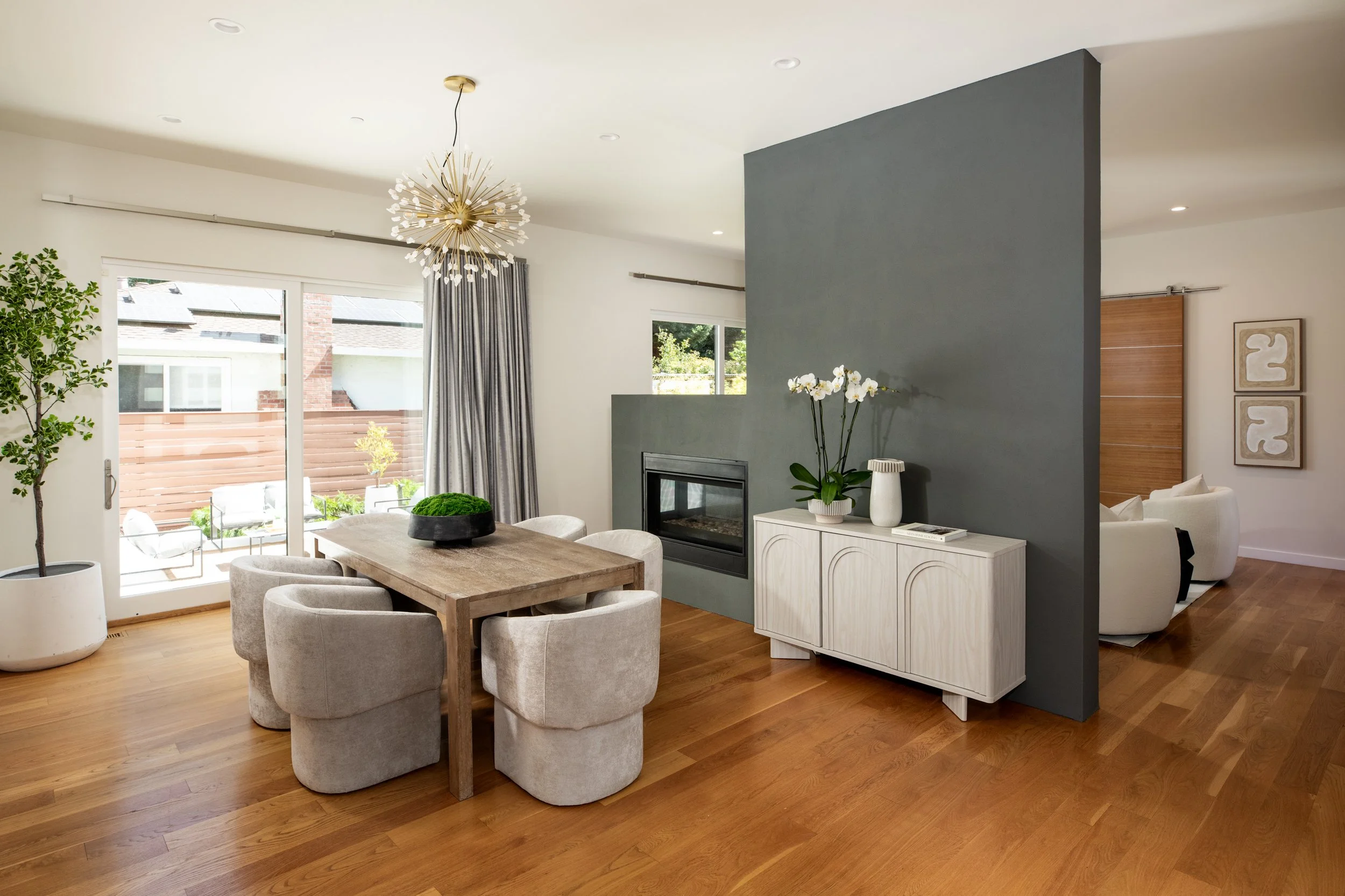 Living room with wooden floor, beige chairs around a wooden dining table, potted plants, a fireplace, large sliding glass door with gray curtains, and modern decor.