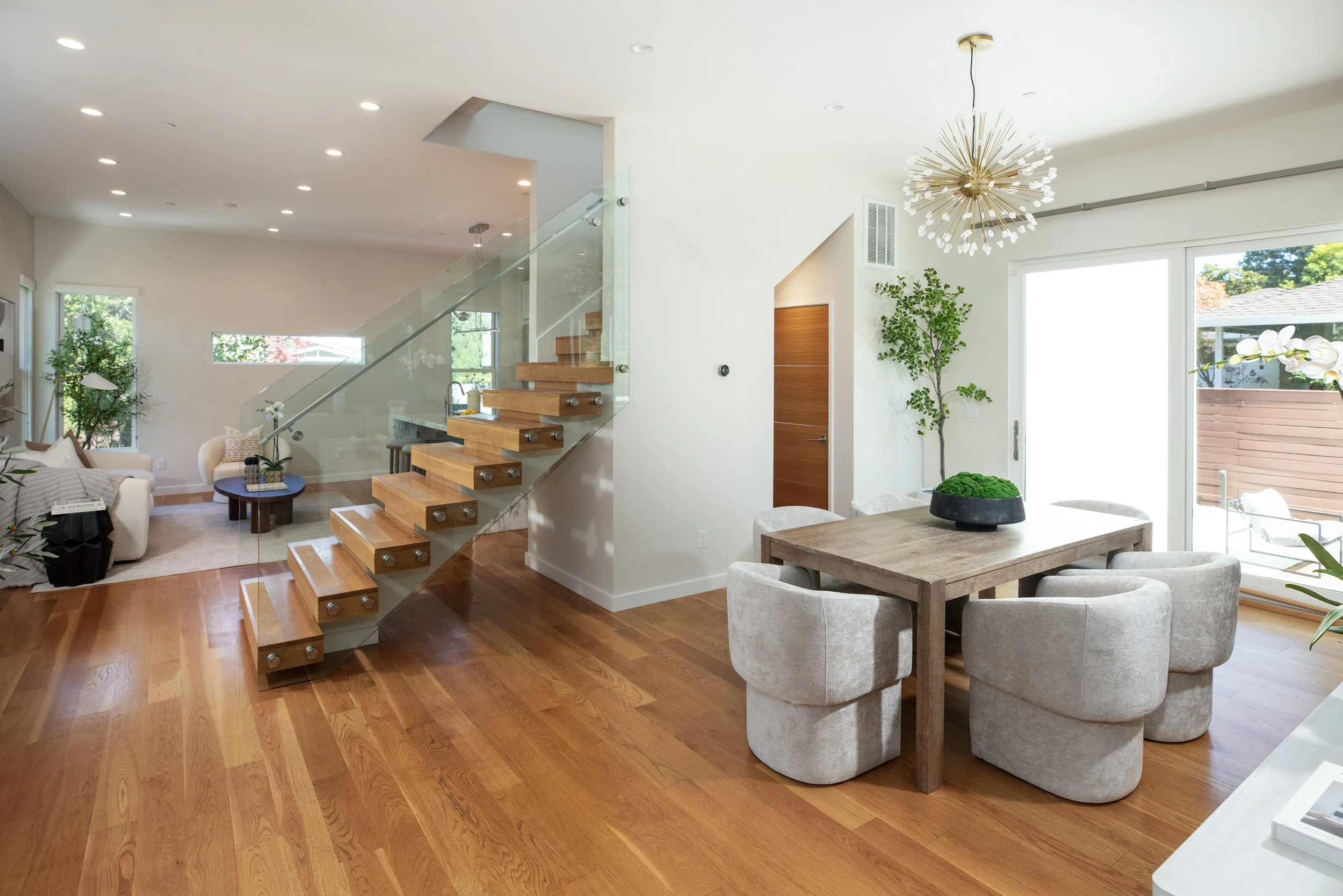 Bright open living and dining area with wooden floors, white and beige furniture, large windows, and modern light fixture.