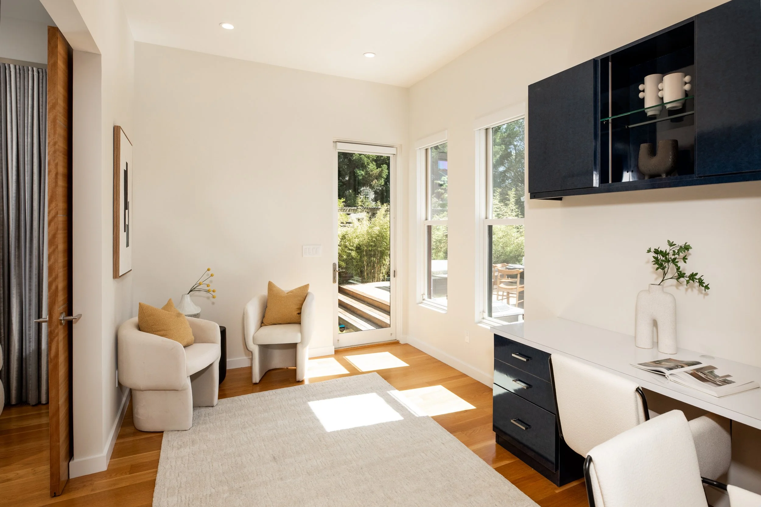 A bright living room with white walls, hardwood flooring, and three windows letting in natural light. There are two white armchairs with tan pillows, a white rug, and a black and white TV cabinet mounted on the wall. A small plant and decorative item