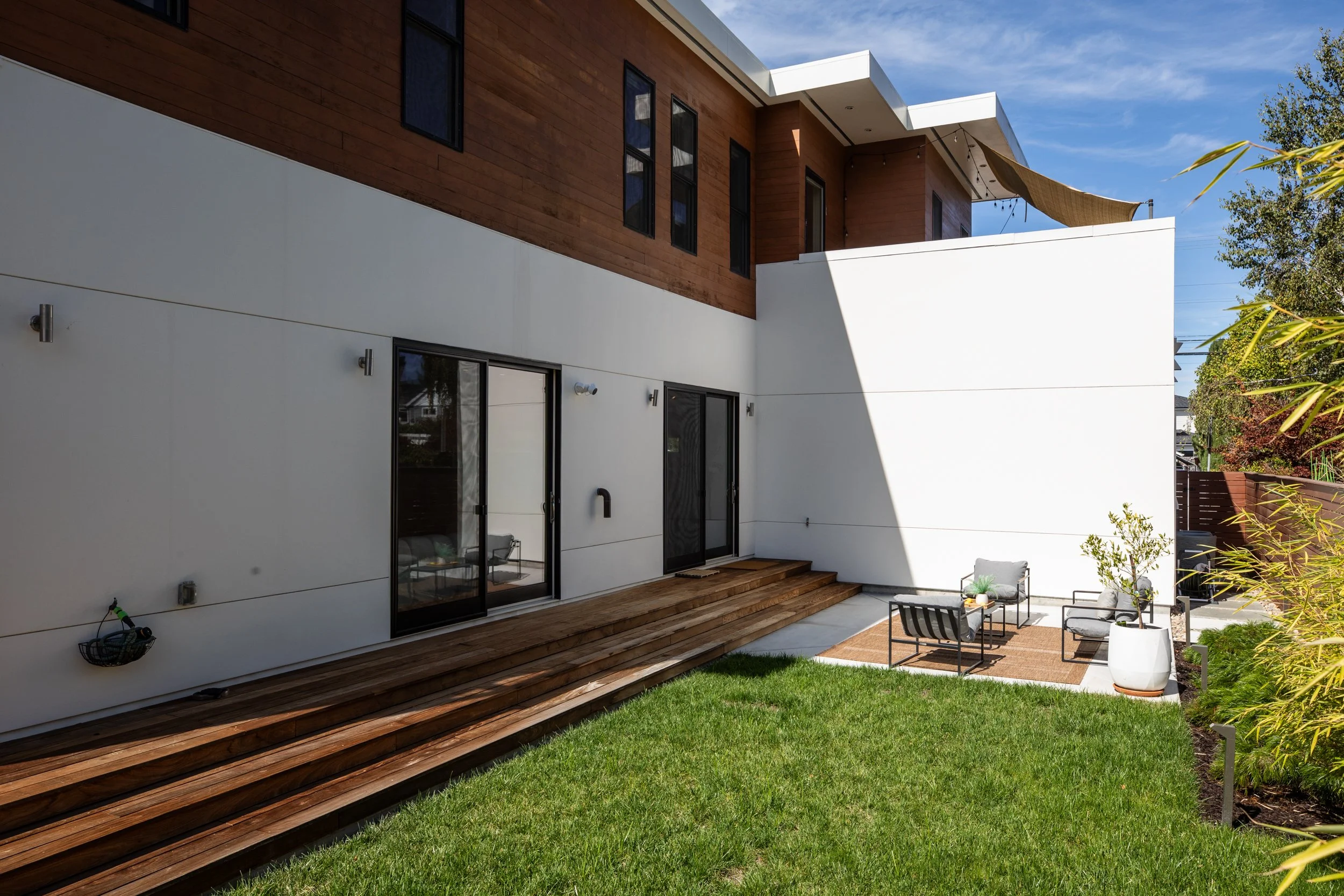 Modern two-story house with white and wooden exterior, glass sliding doors, and a backyard seating area with chairs, a table, and potted plants, under a partly cloudy sky.