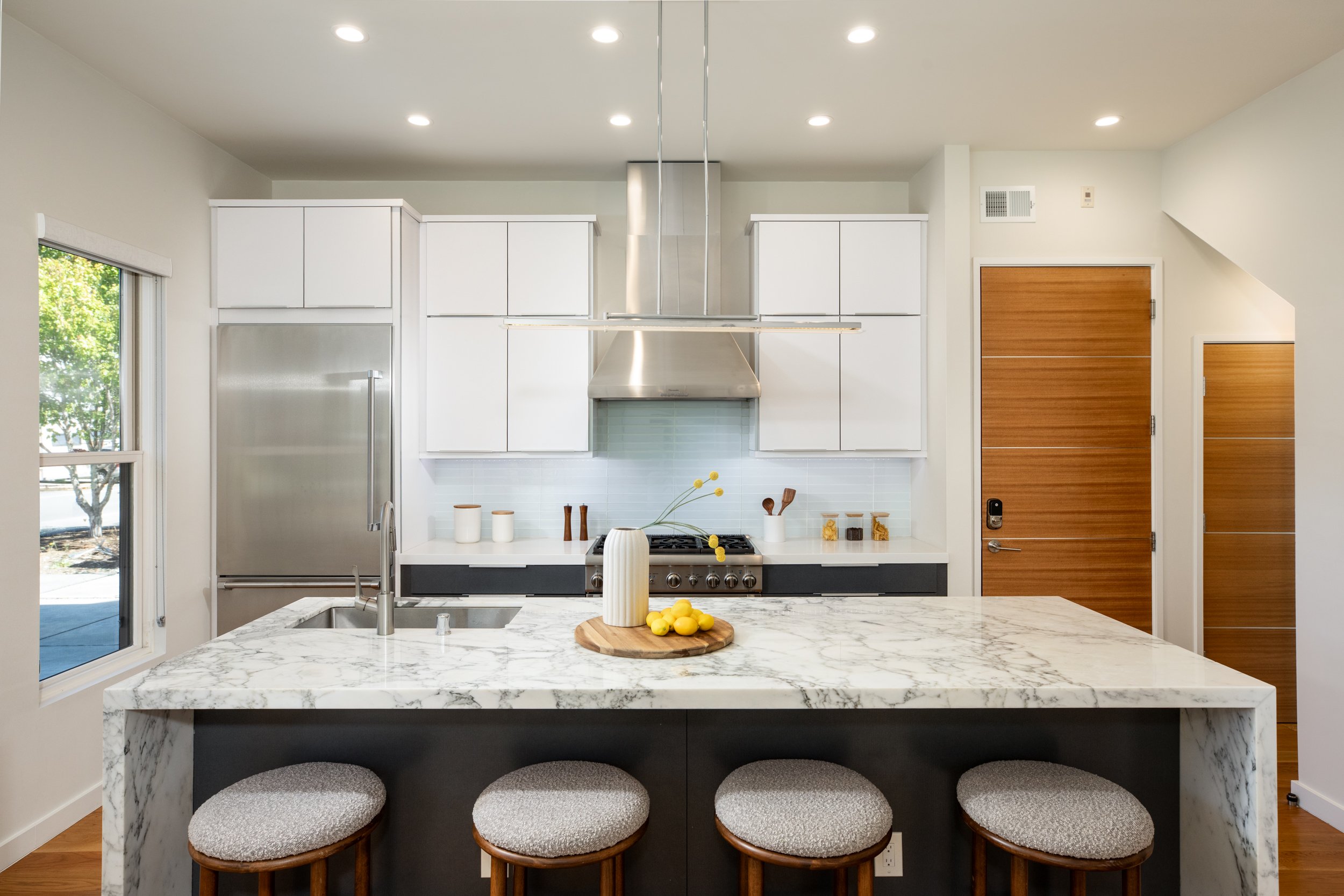 Modern kitchen with white cabinets, stainless steel refrigerator, marble island with three cushioned stools, and wooden doors.