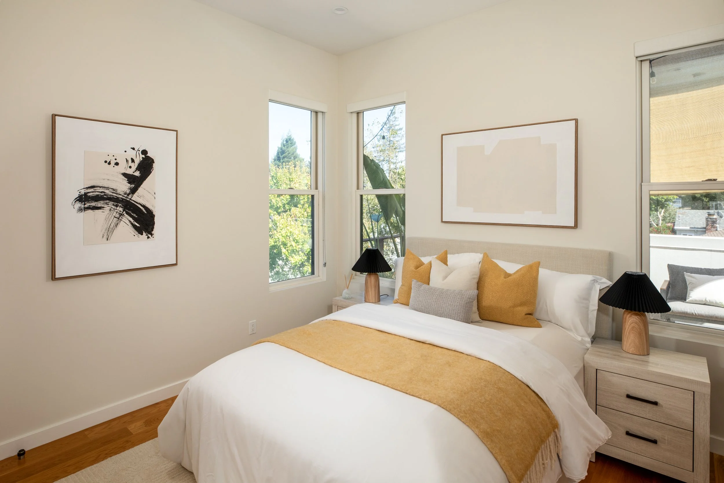 A cozy bedroom with two large windows showing greenery outside, beige walls, a bed with white bedding and various yellow and gray pillows, wooden nightstands with black lamps, and framed abstract art on the walls.