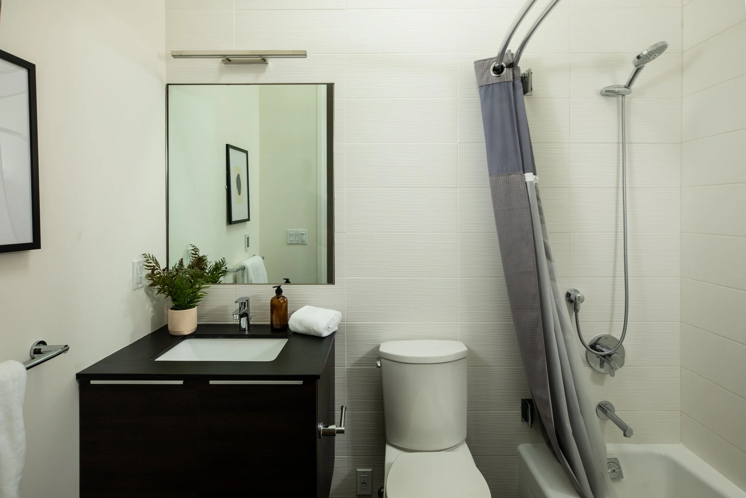 A modern bathroom with a white wall and a large mirror above a black vanity with a sink. On the vanity, there is a potted plant, a soap dispenser, a towel, and a folded towel. To the right, there is a wall-mounted toilet and a shower with a gray curt