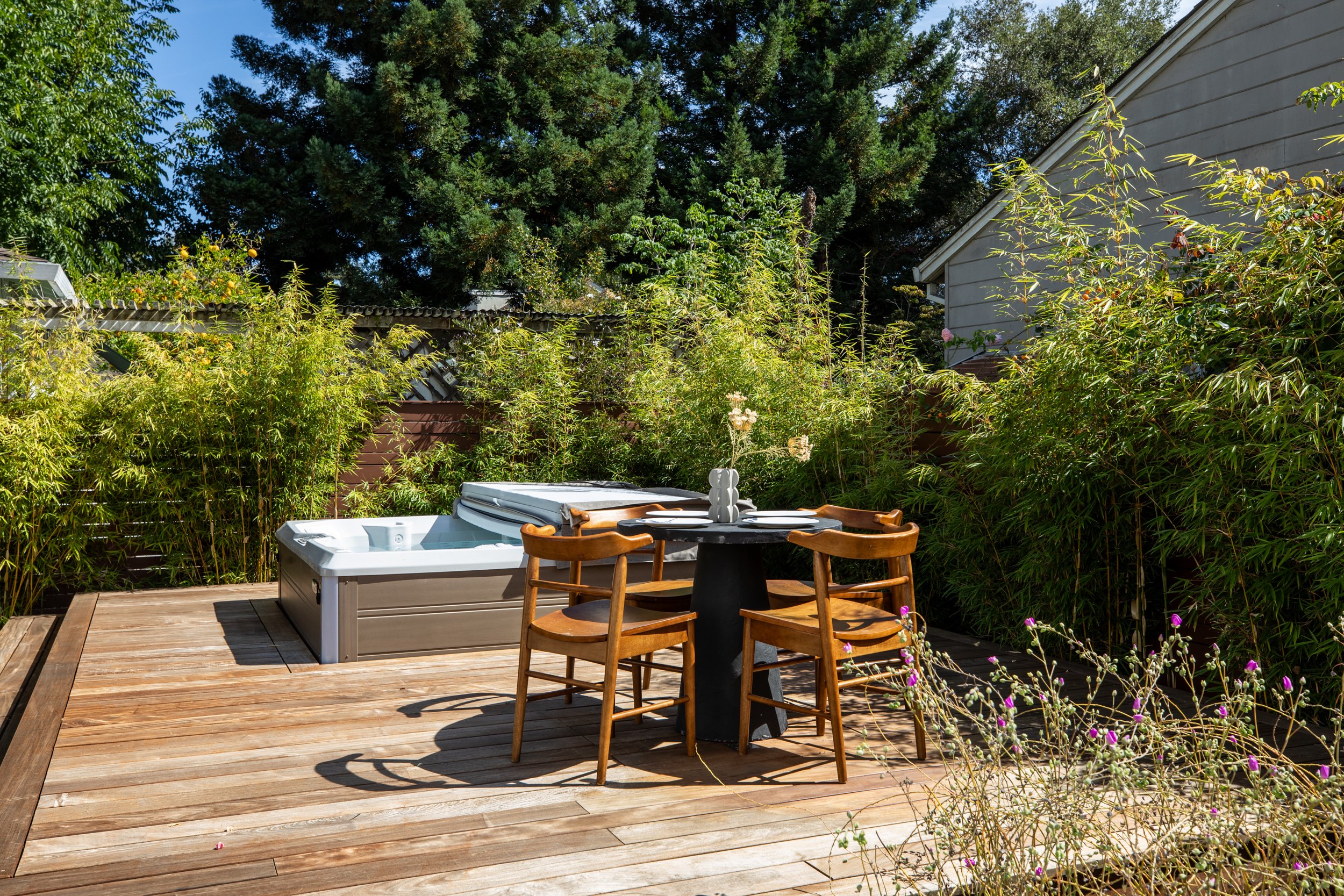 Outdoor patio with wooden deck furniture, including a round black table with four wooden chairs, a hot tub, and lush green trees and bushes surrounding the area.